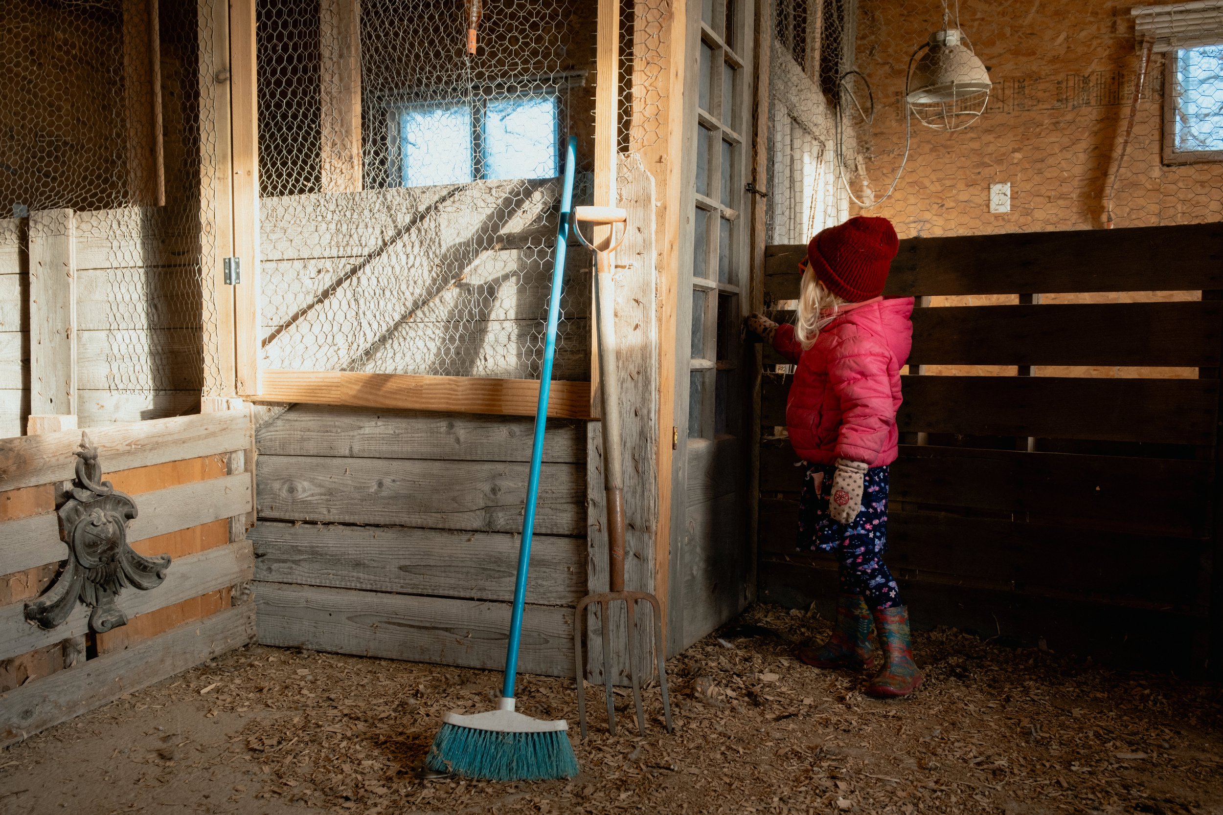 A young girl with long blonde hair, wearing a red knit hat, pink jacket, patterned gloves, and flowered boots, standing inside a rustic barn with wooden walls and dirt floor, holding a farming tool, with a broom and rake leaning nearby.