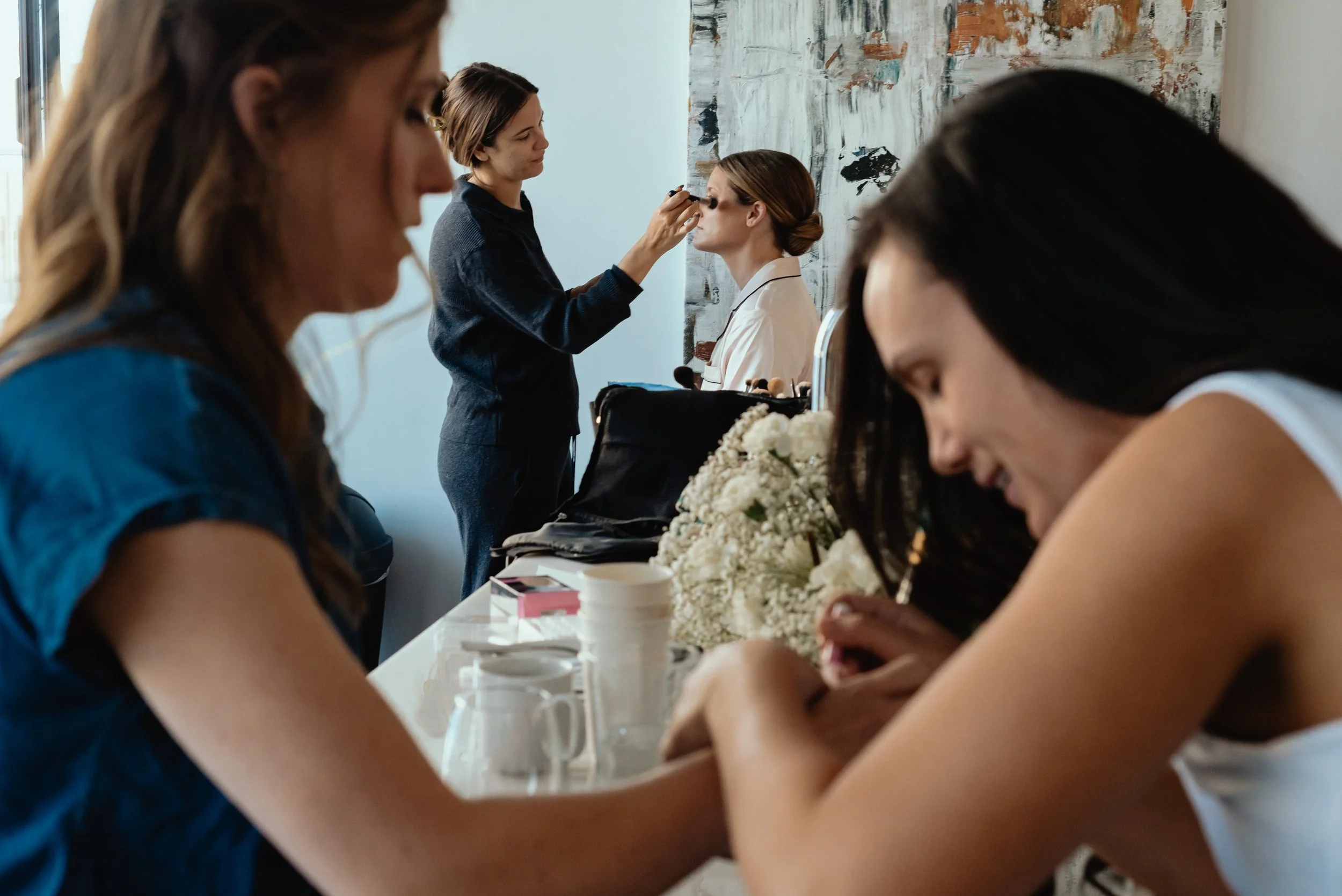 A candid, documentary-style shot of a bride getting her makeup done, framed by a soft, out-of-focus foreground of bridesmaids adjusting wrist jewelry.