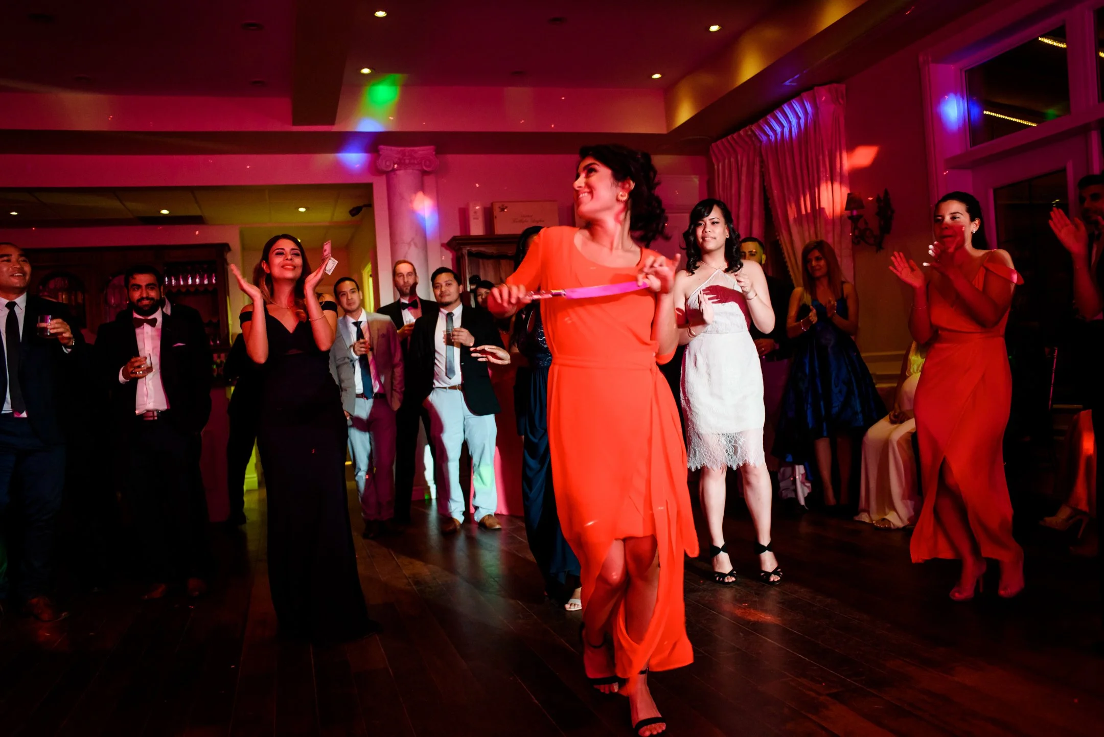 Women in formal attire celebrating at a party with colorful lights and a dance floor. A woman in red dress is performing the Persian knife dance, known as Raghseh Chagoo.