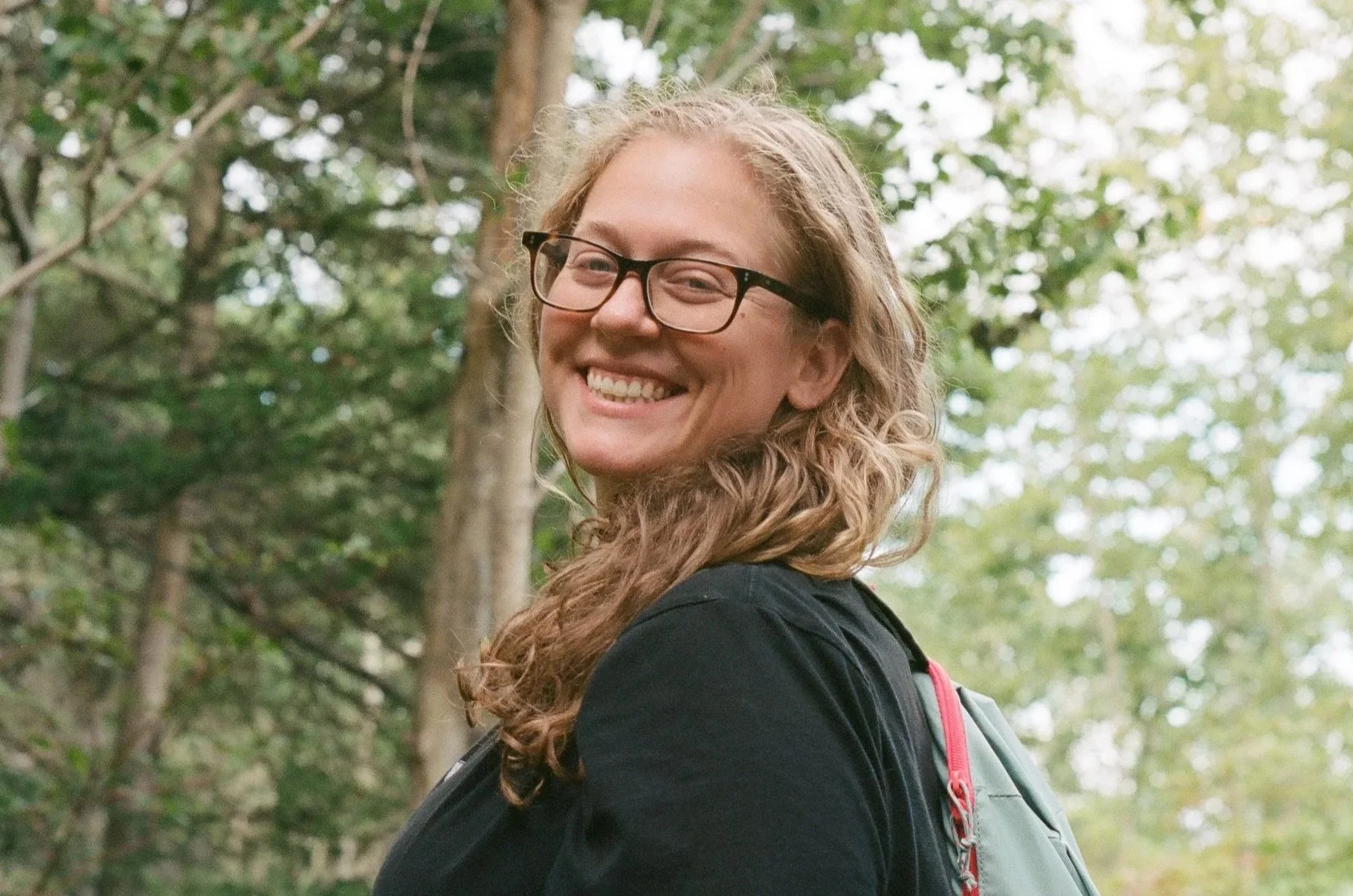A young woman with curly light blonde hair, wearing glasses and a black shirt, smiling and looking back over her shoulder outdoors in a forested area with trees and green foliage.