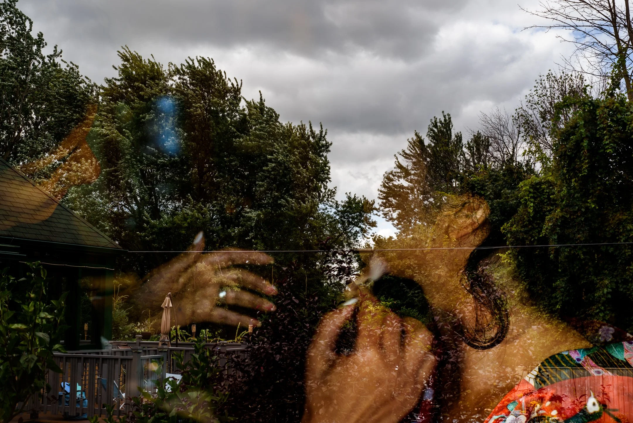 A backyard with trees, cloudy sky, and a fence, with some reflections or superimposed images of a person getting makeup applied