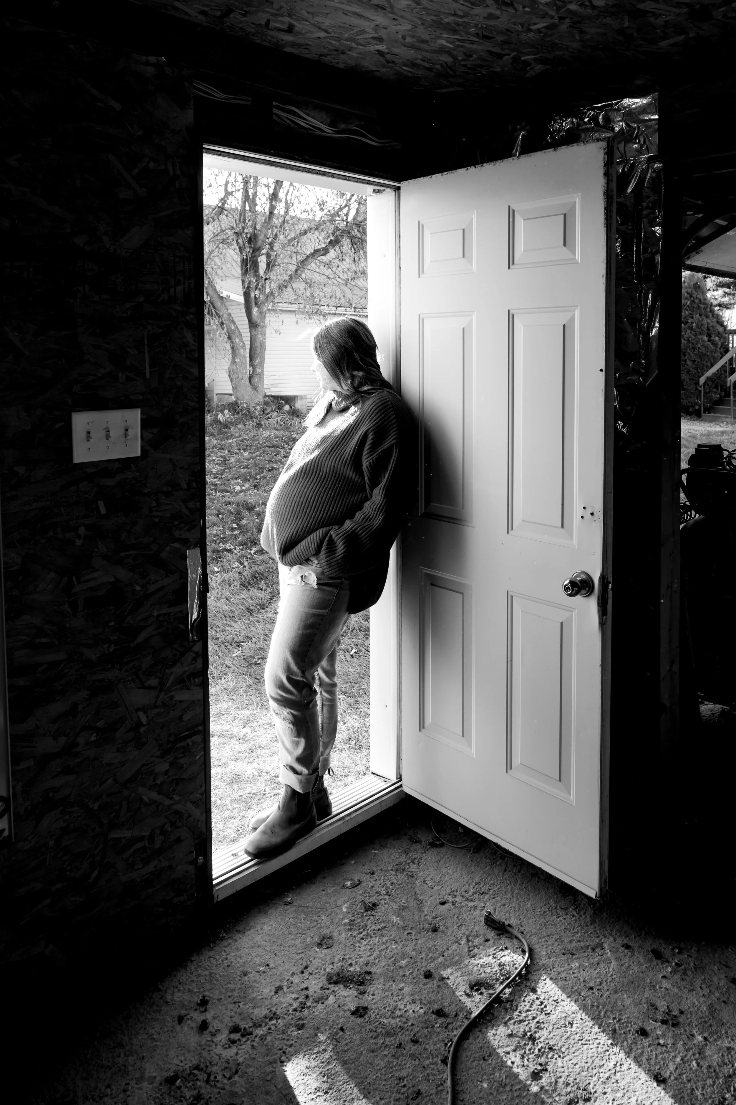 A pregnang woman standing at the open door of a building, looking outside into a yard with a tree and a house in the background, in black and white.