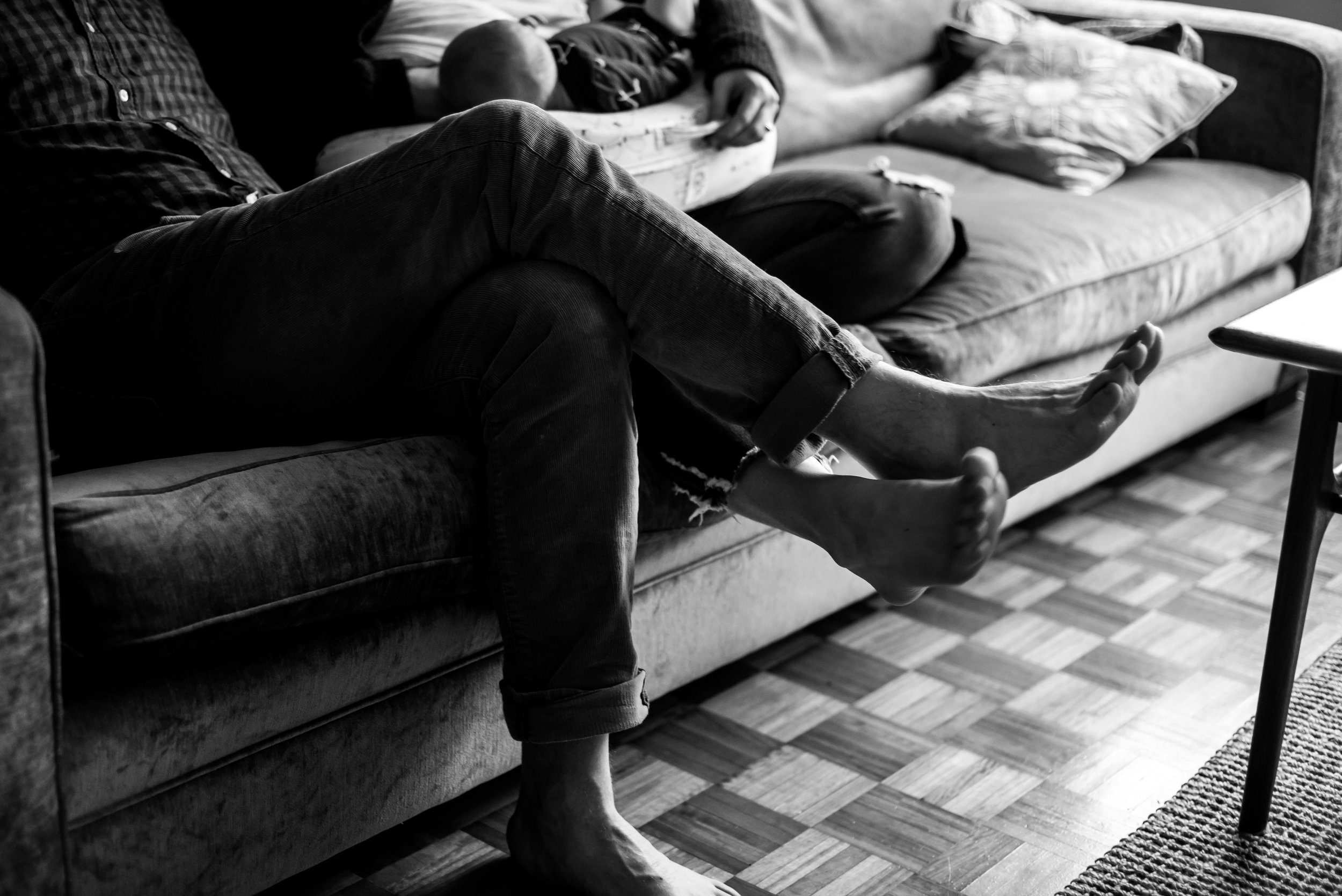 Black and white photo of a couple holding a baby vintage couch with pillows in a cozy living room, with patterned wooden flooring visible.