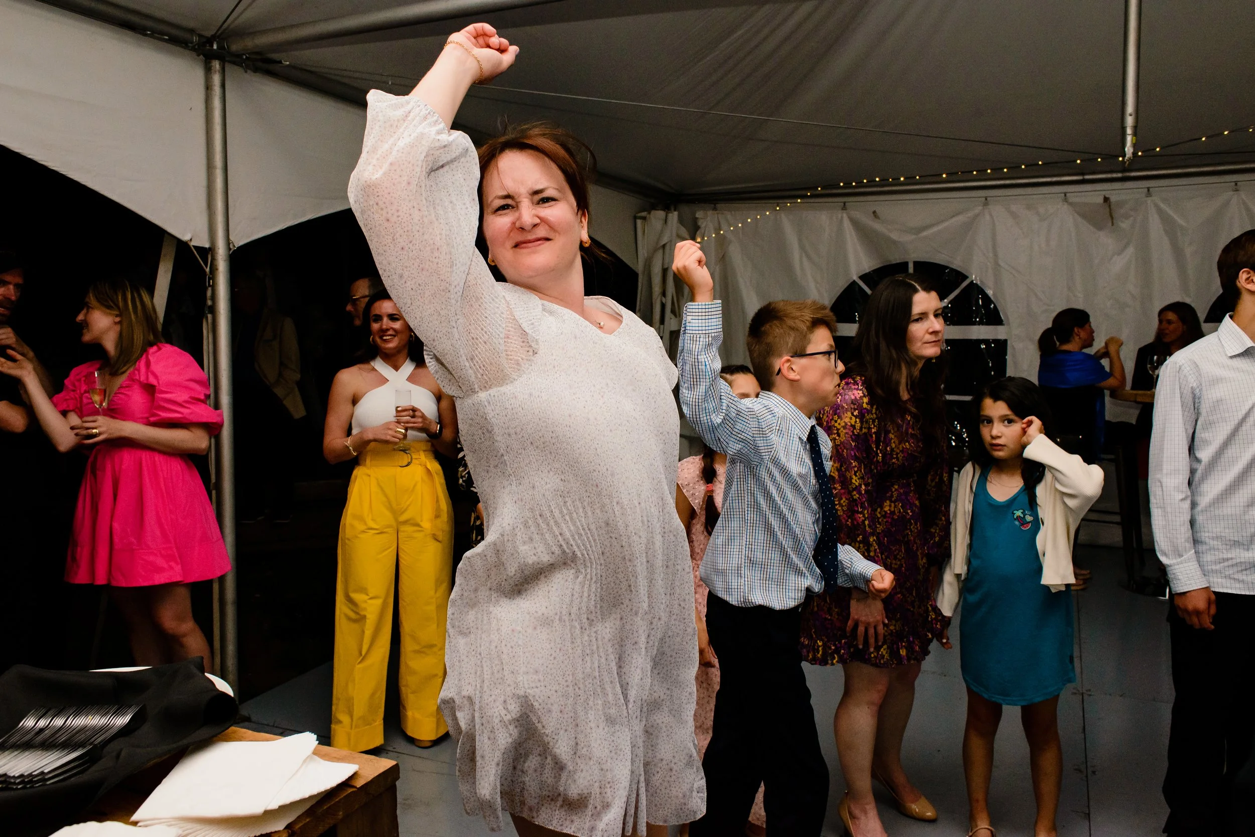 Wedding guests dancing in tent focused on woman guest jumping with raised arm look at the camera.