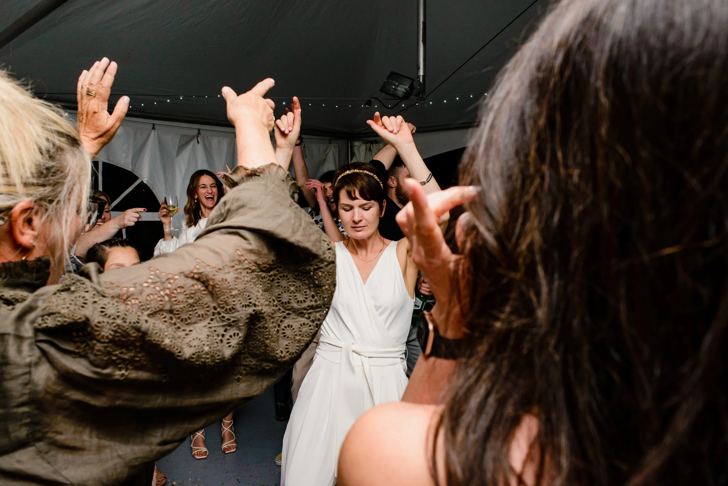 Wedding guests dancing in tent cheering on bride in white pantsuit dancing with raised hands and closed eyes