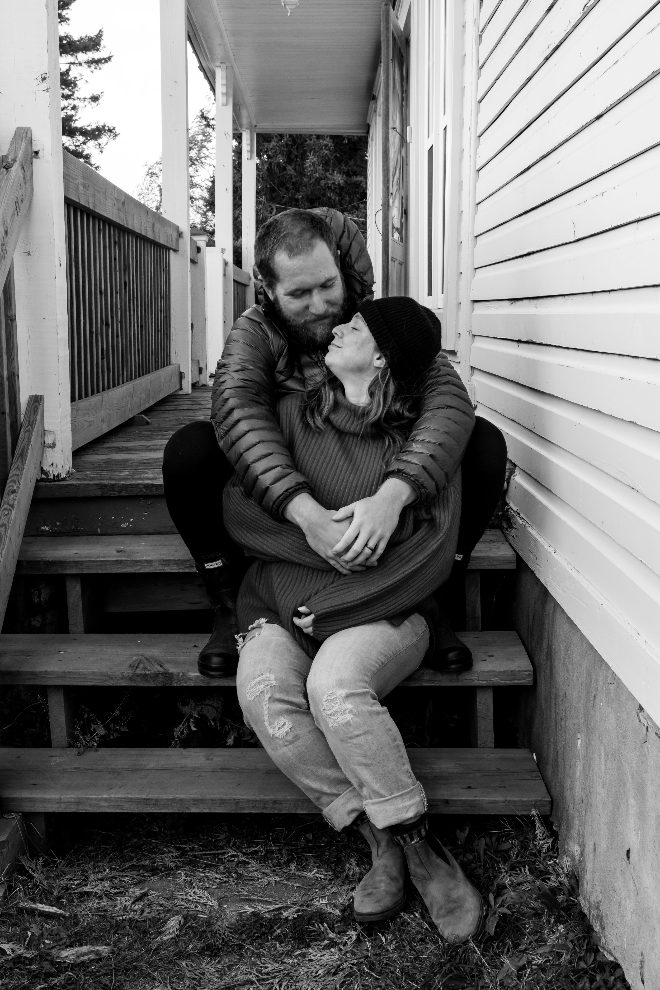 A couple sitting close together on wooden stairs outside a house. The man is leaning over the woman, and they are looking into each other's eyes with affection. The woman has long hair and is wearing a beanie and a sweater. The man has a beard and is