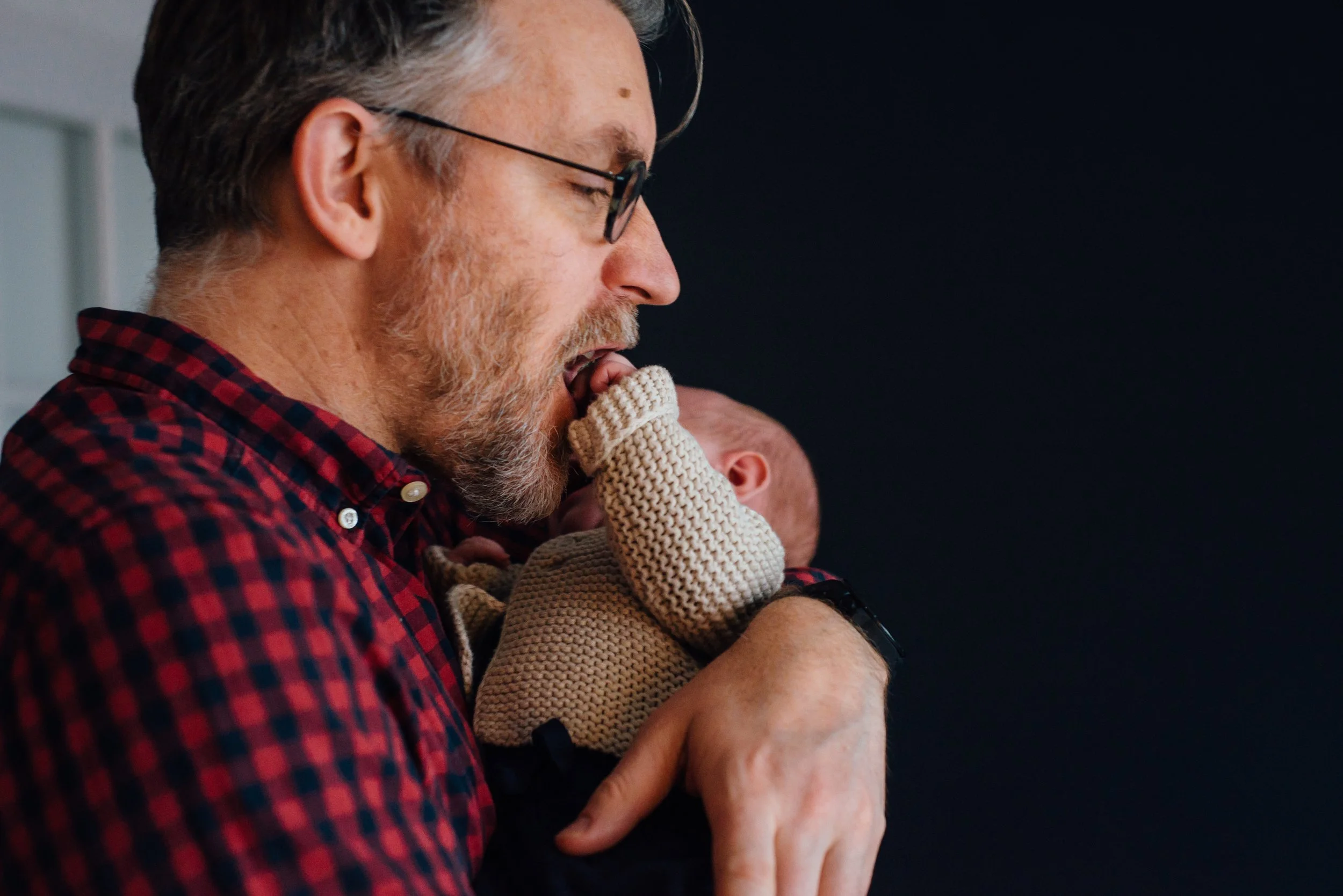 A man with glasses and a beard, wearing a red and black checkered shirt, with eyes closed, gently holding a baby's legs. The baby is wearing a beige sweater and is partially visible. The background is dark.