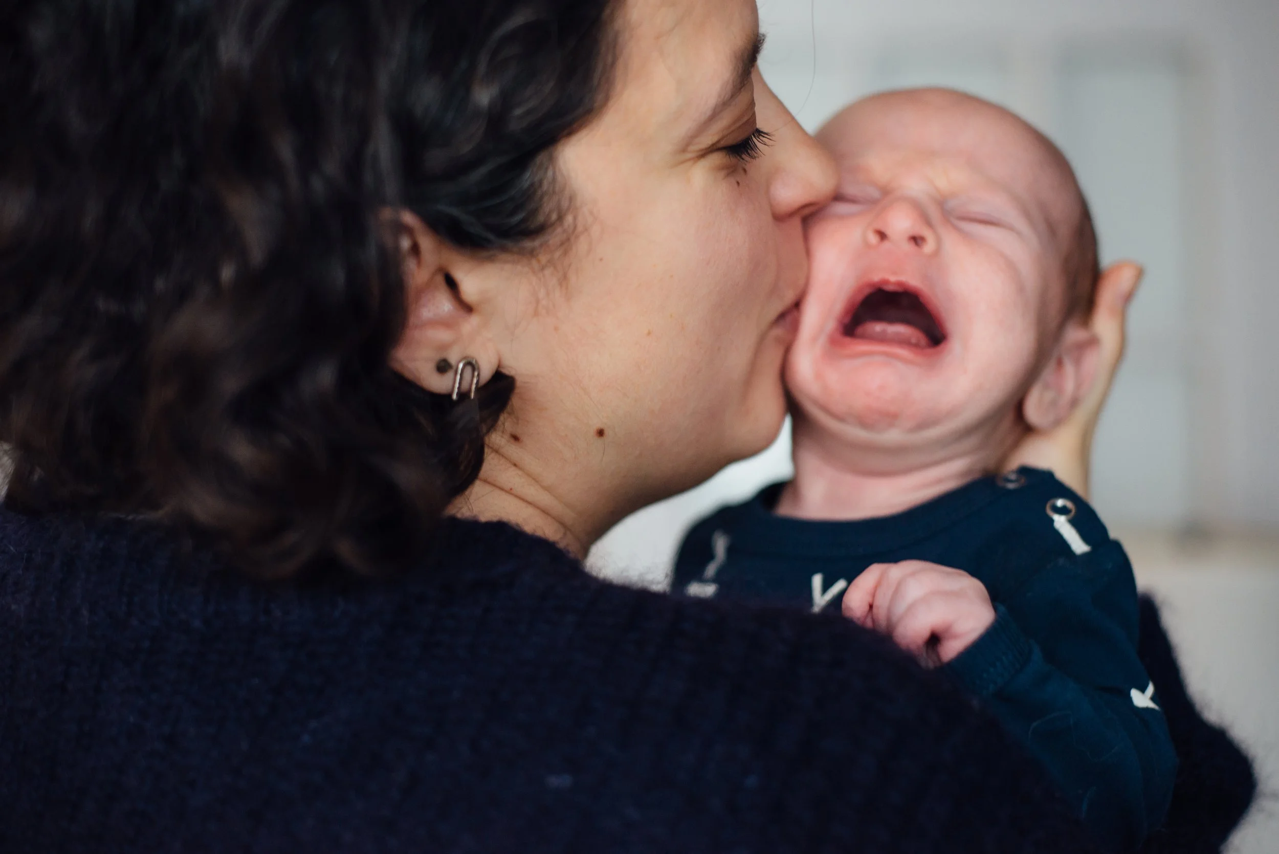 A woman with dark, curly hair and earrings is holding a crying baby close to her face.