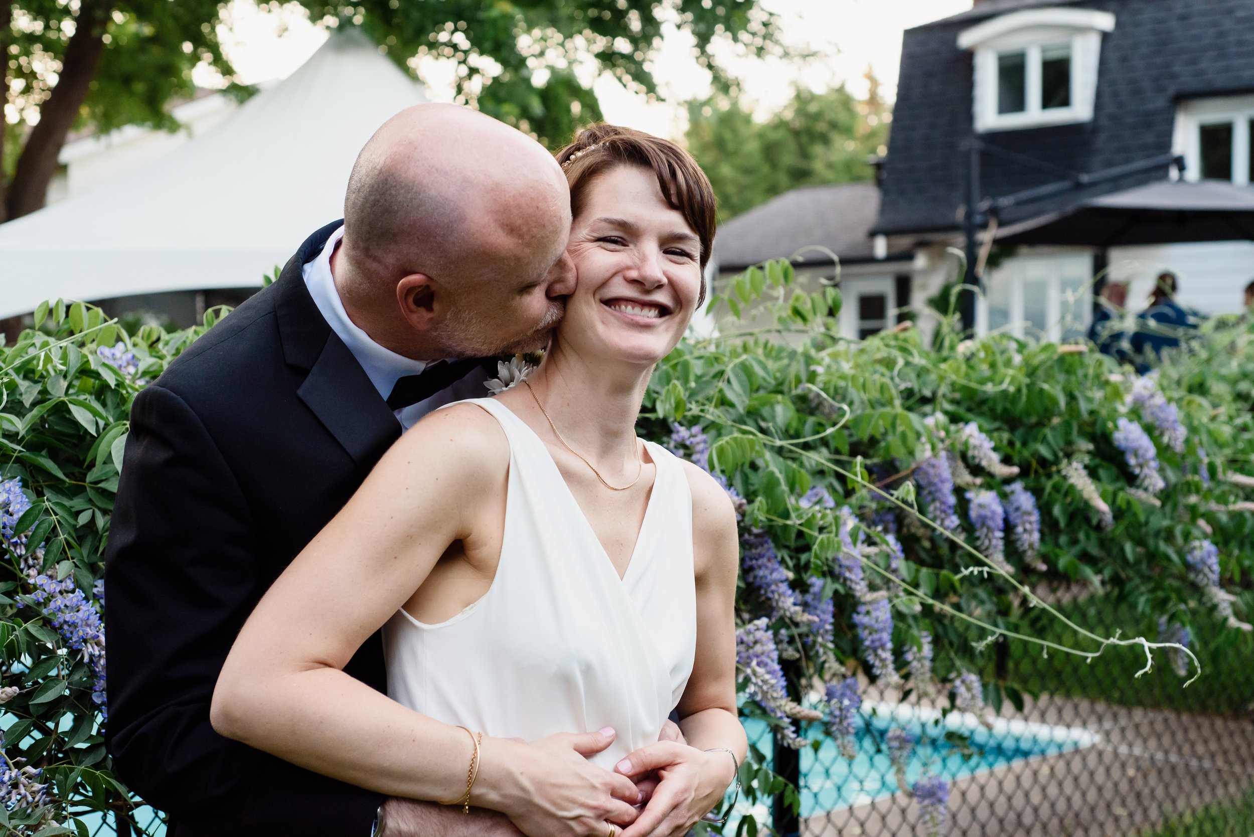 Closeup of couple embracing in front of wisteria on fenced in pool. Bride smiles to camera as groom kisses her cheek.