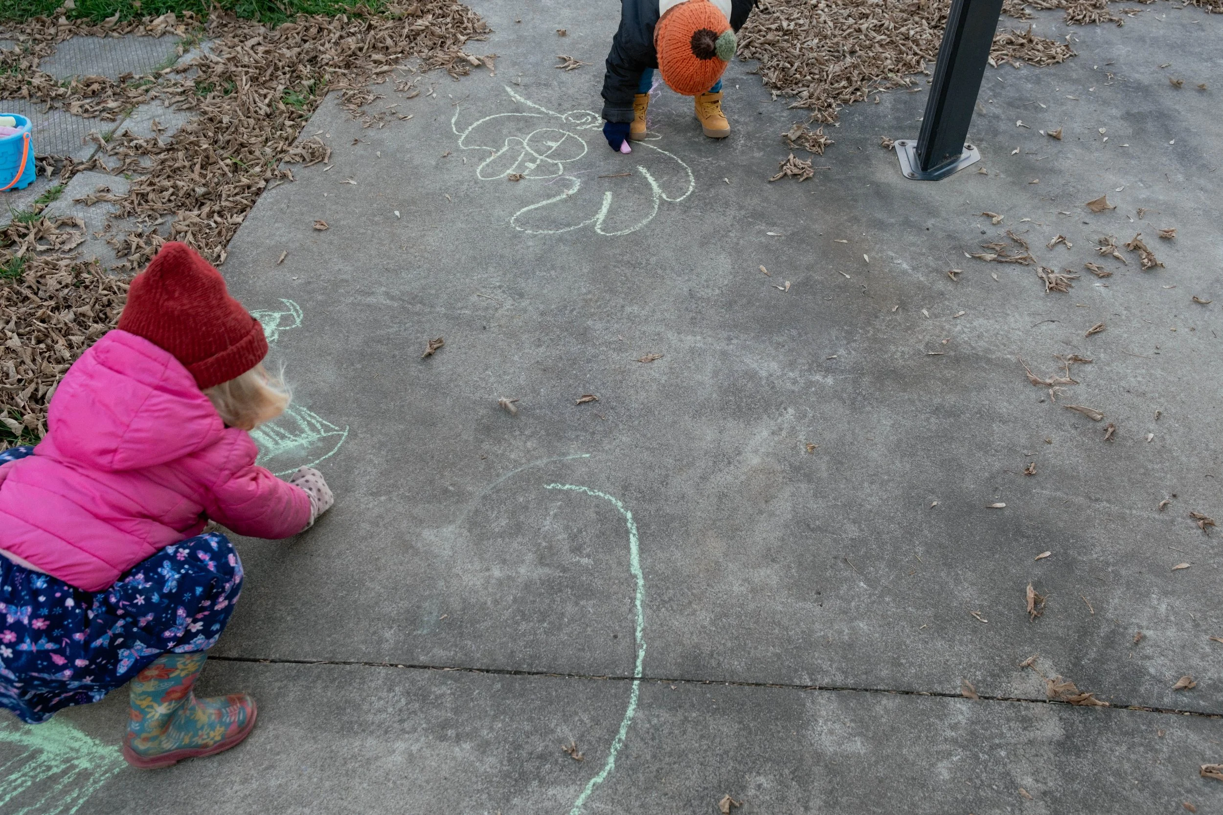 Two children drawing with chalk on a concrete sidewalk, surrounded by fallen leaves. One child is wearing a pink jacket, red hat, and patterned pants, and the other child is wearing a dark jacket and brown boots with a pumpkin knit hat.