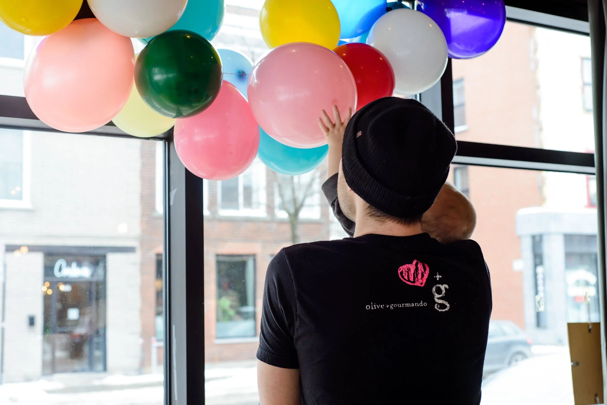 A person wearing a black beanie and a black shirt with pink and white text, holding a baby, is reaching towards a cluster of colorful balloons inside a building near large windows.