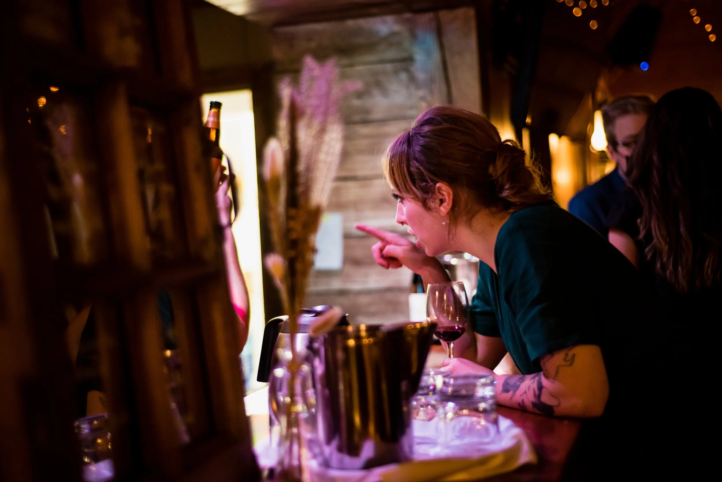 A woman inside a cozy, dimly lit bar or restaurant, leaning over a table, pointing at something, with a glass of red wine in front of her. The background has wooden walls and soft lighting, with other patrons partially visible.