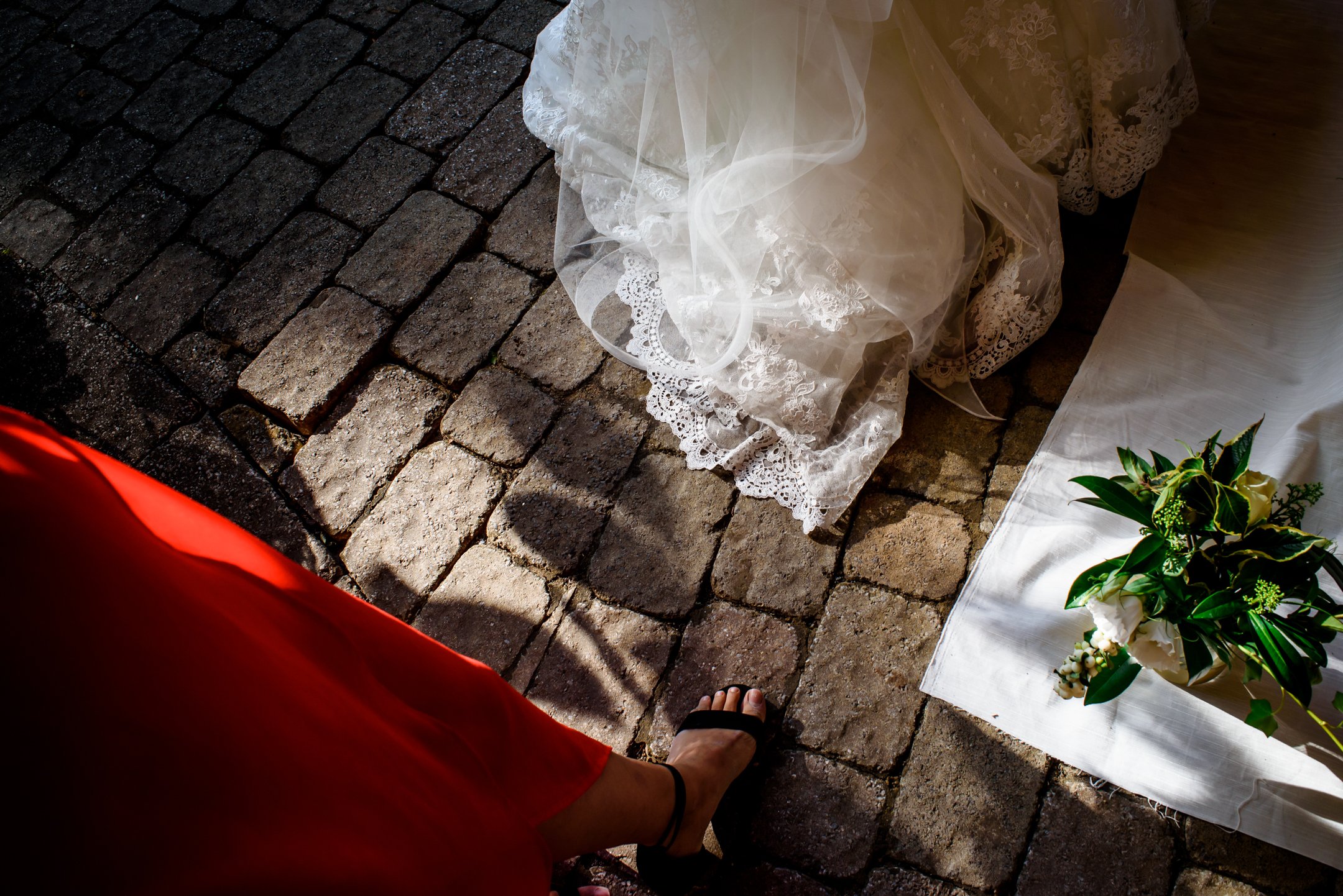 A person wearing a red dress and black sandals standing on a cobblestone street, next to a small table with a white lace tablecloth, a bouquet of white flowers with greenery, and a sheer lace veil.