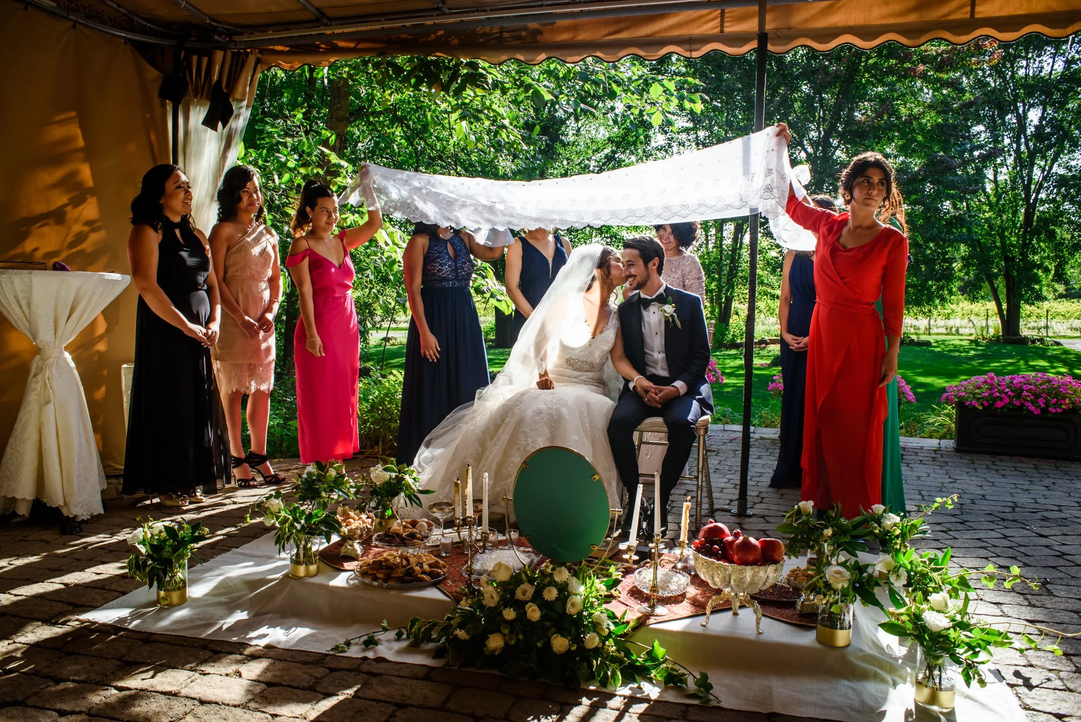 A persian wedding ceremony outdoors with a bride and groom sitting under a white cloth, surrounded by bridesmaids and guests. In front of them sits the traditional table of offerings, Sofreh Aghd.