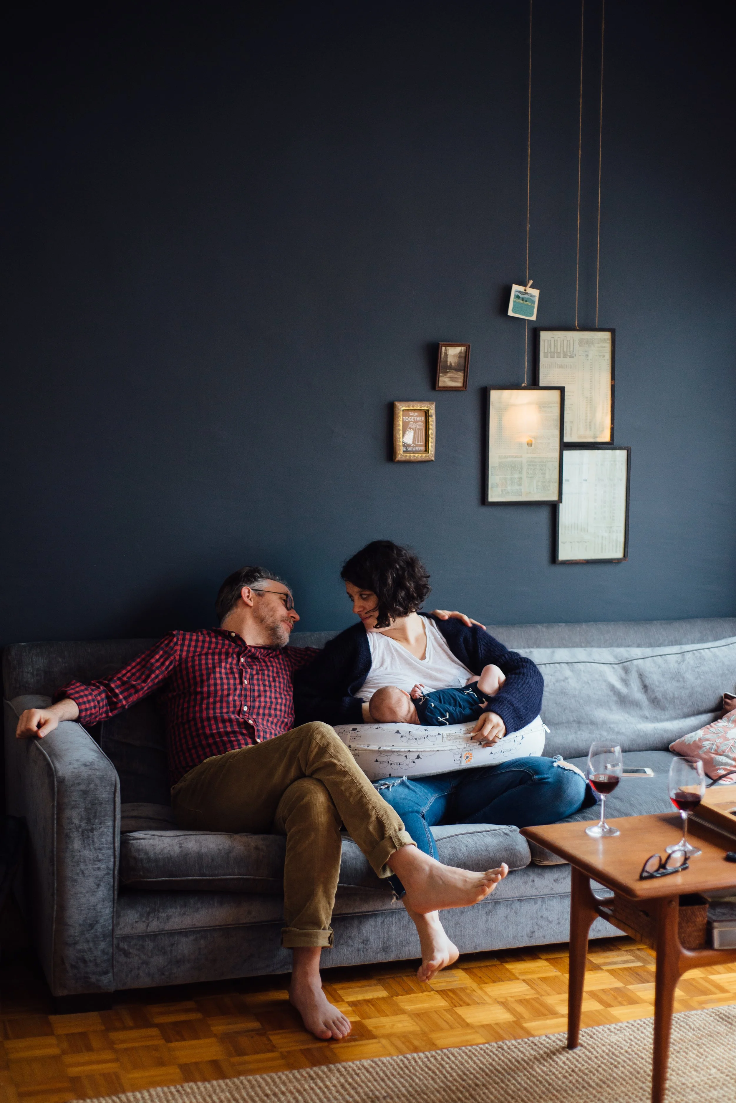 A man and woman sit closely on a dark gray sofa, sharing an intimate moment. The woman has curly hair and is holding a baby, while the man, wearing glasses and a red plaid shirt, looks at her. There are glasses of wine, a phone, and a pair of glasses