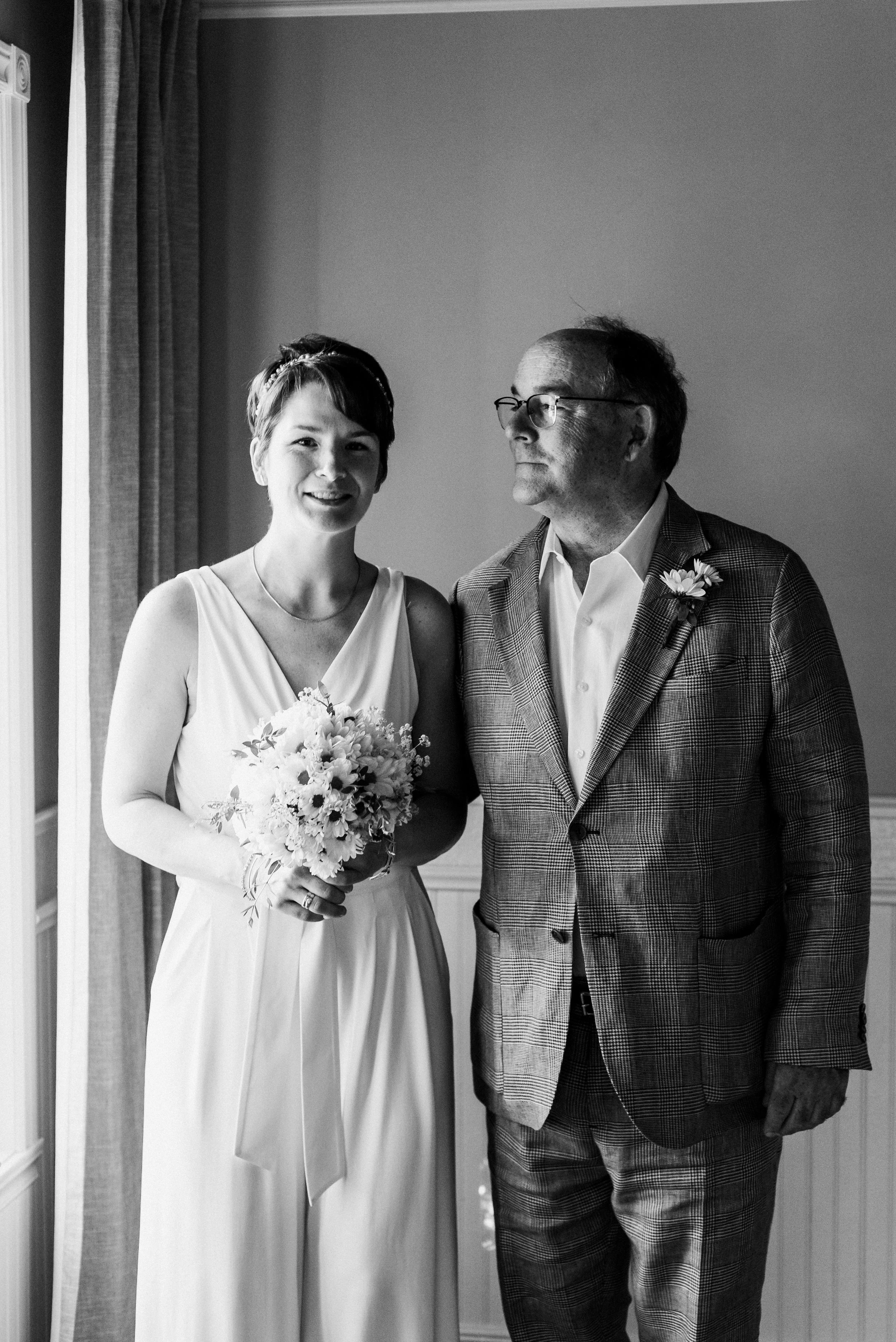 Black and white (BW) photo of bride standing next to father. Bride staring strait into camera as father looks over to her. 