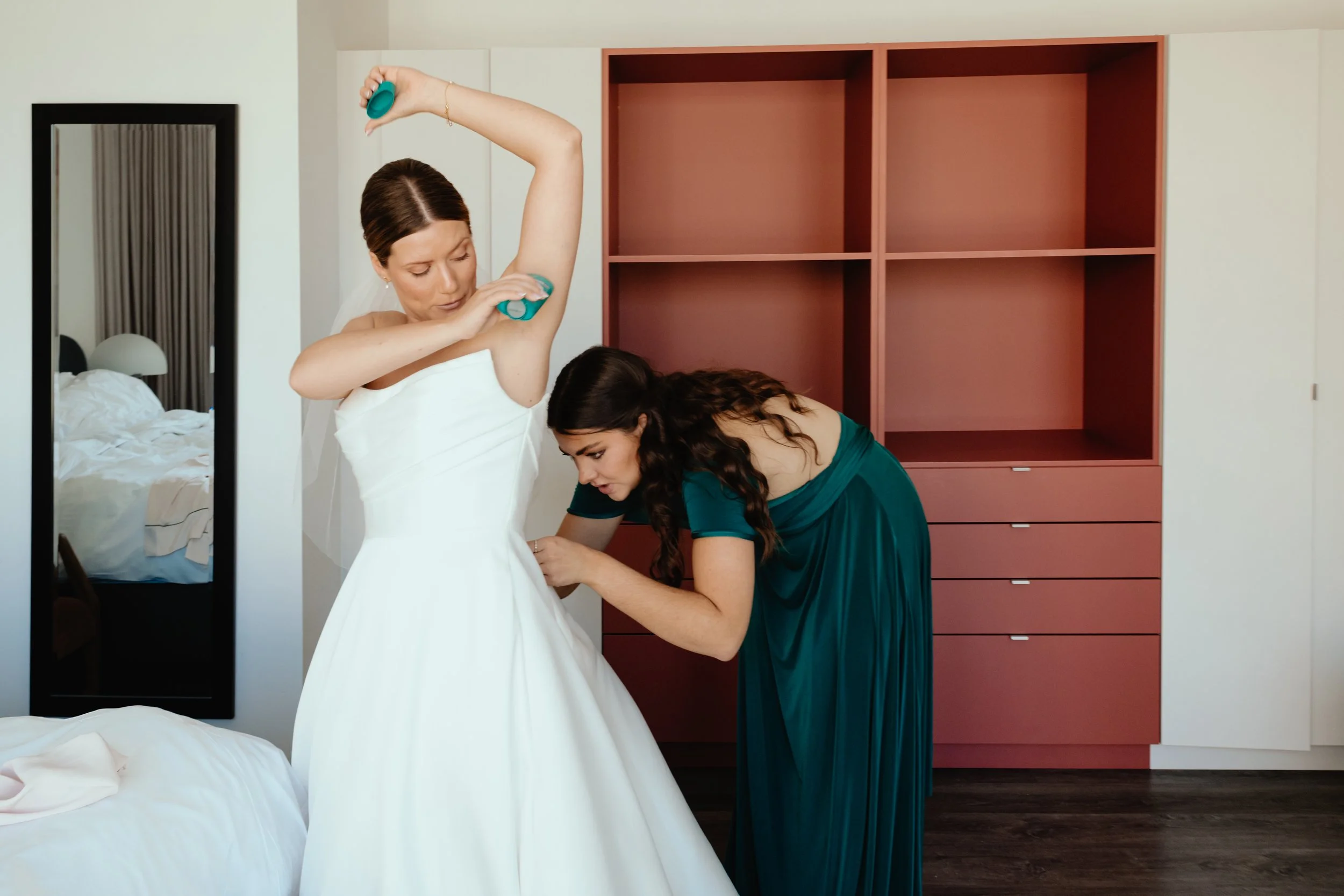 A candid, real-life photograph captures a raw and humorous behind-the-scenes moment of wedding day preparation. In the Old Montreal bridal suite, a bride casually applies deodorant while her bridesmaid works on buttoning up the back of her wedding dr