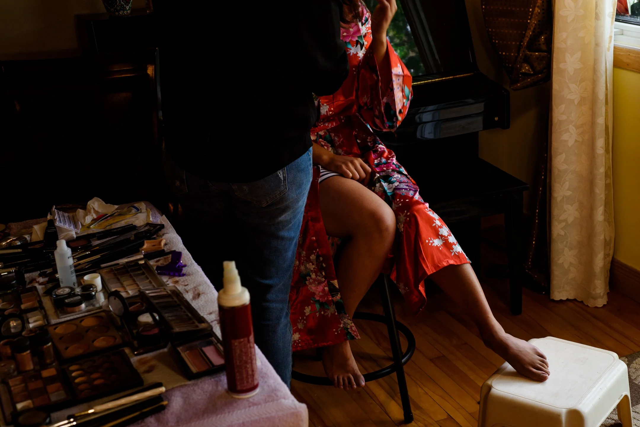 A woman in a red floral kimono sitting on a chair with her leg raised, having her makeup done by another person. Makeup supplies and tools are on a table nearby, and a petticoat is on the floor.
