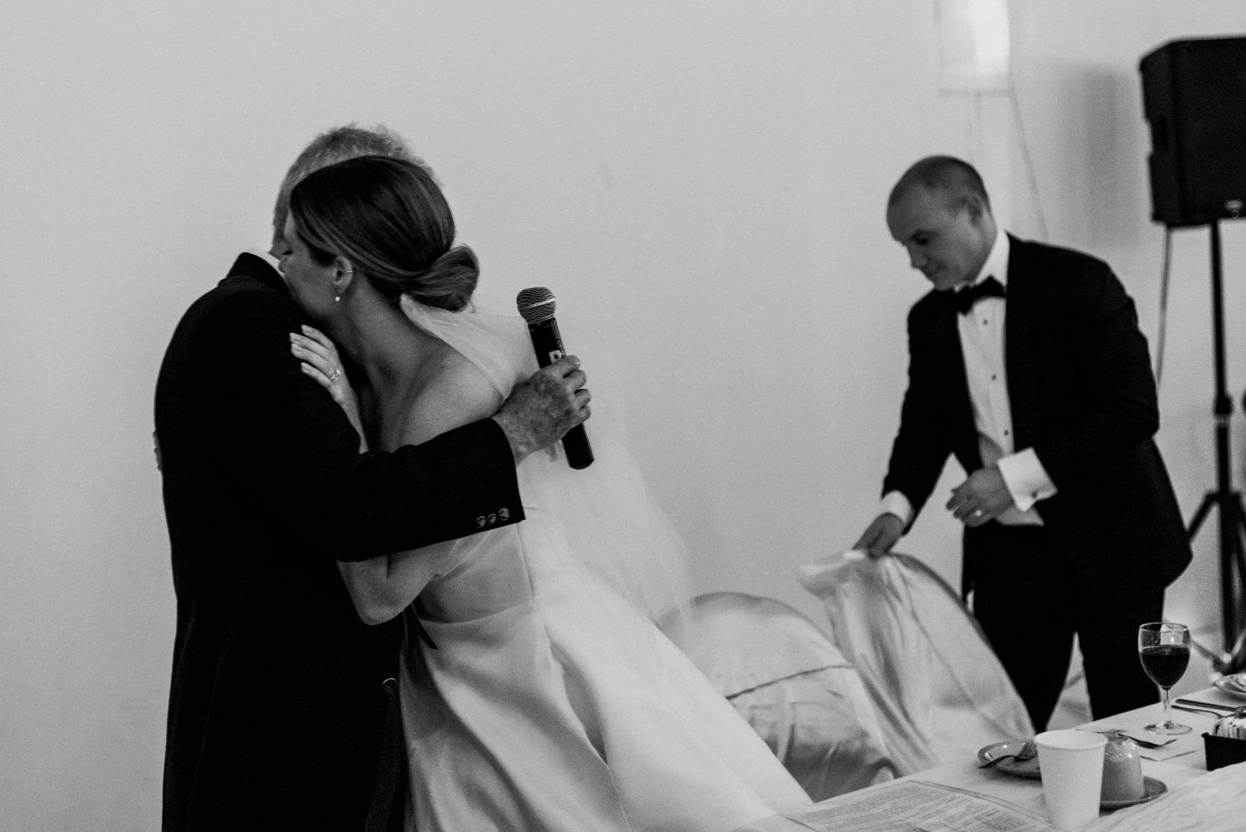 Black and white wedding photo of a bride embracing her father after his speech, as he holds a microphone and the groom gets up from his seat in the background.