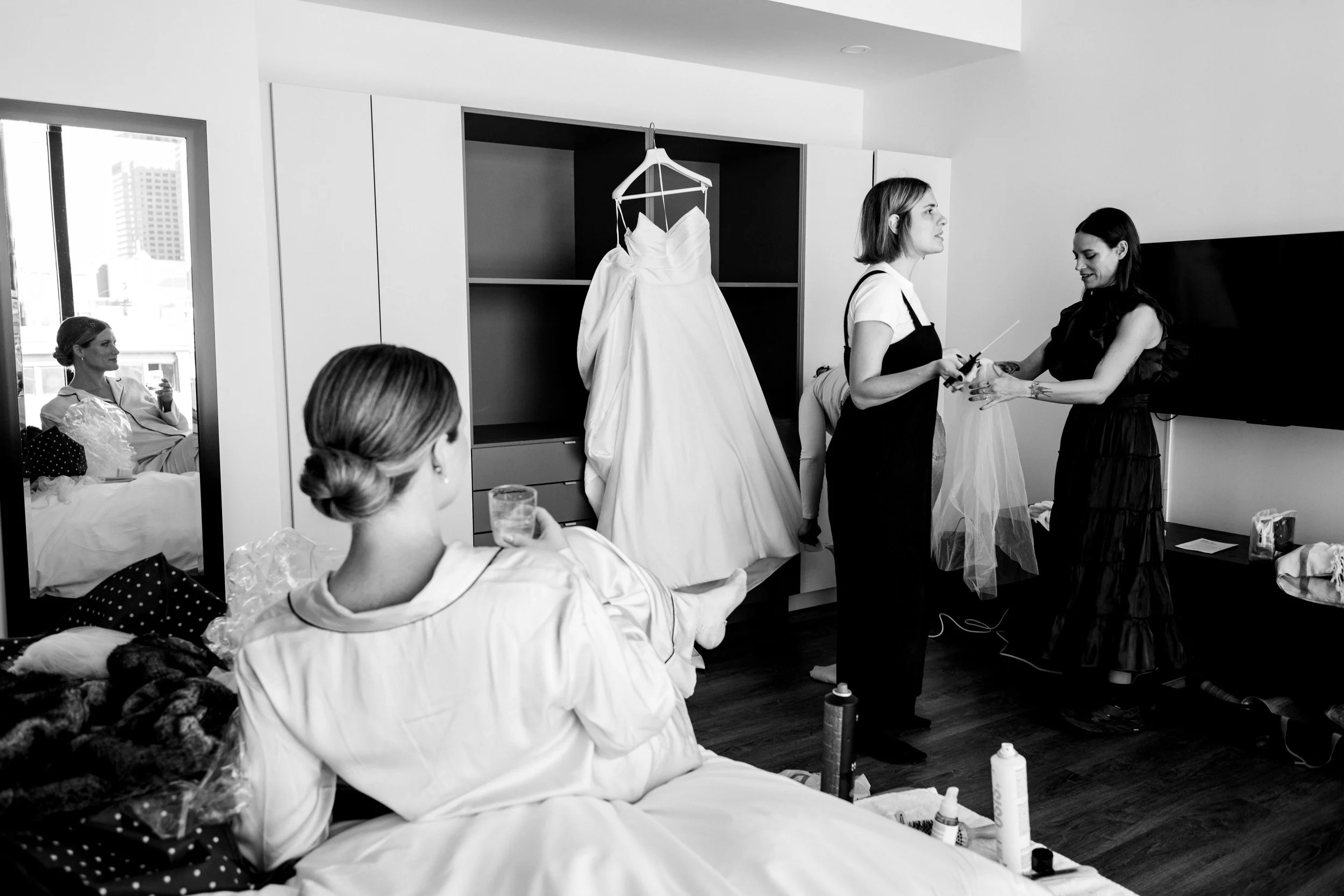 A joyful black-and-white photograph inside a bridal suite, featuring a bride in sil pajamas seated in the foreground. Her reflection is visible in a mirror as she smiles and holds a champagne flute. In the background, the dress hangs proudly as bride