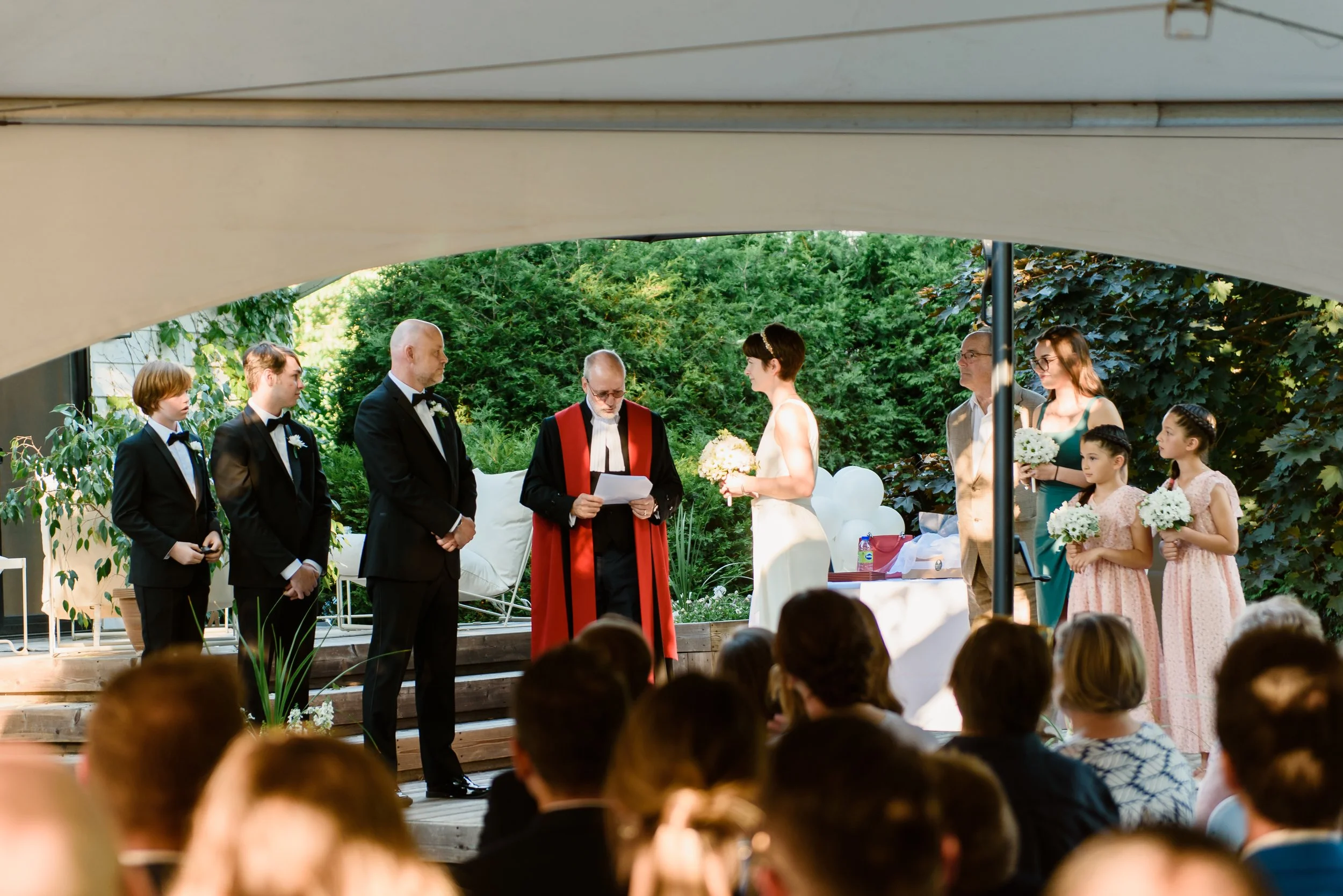 An outdoor backyard wedding ceremony with a celebrant wearing full judge robes in black and red with a white collar. The celebrant stands at the altar, positioned between the wedding couple and the wedding party.