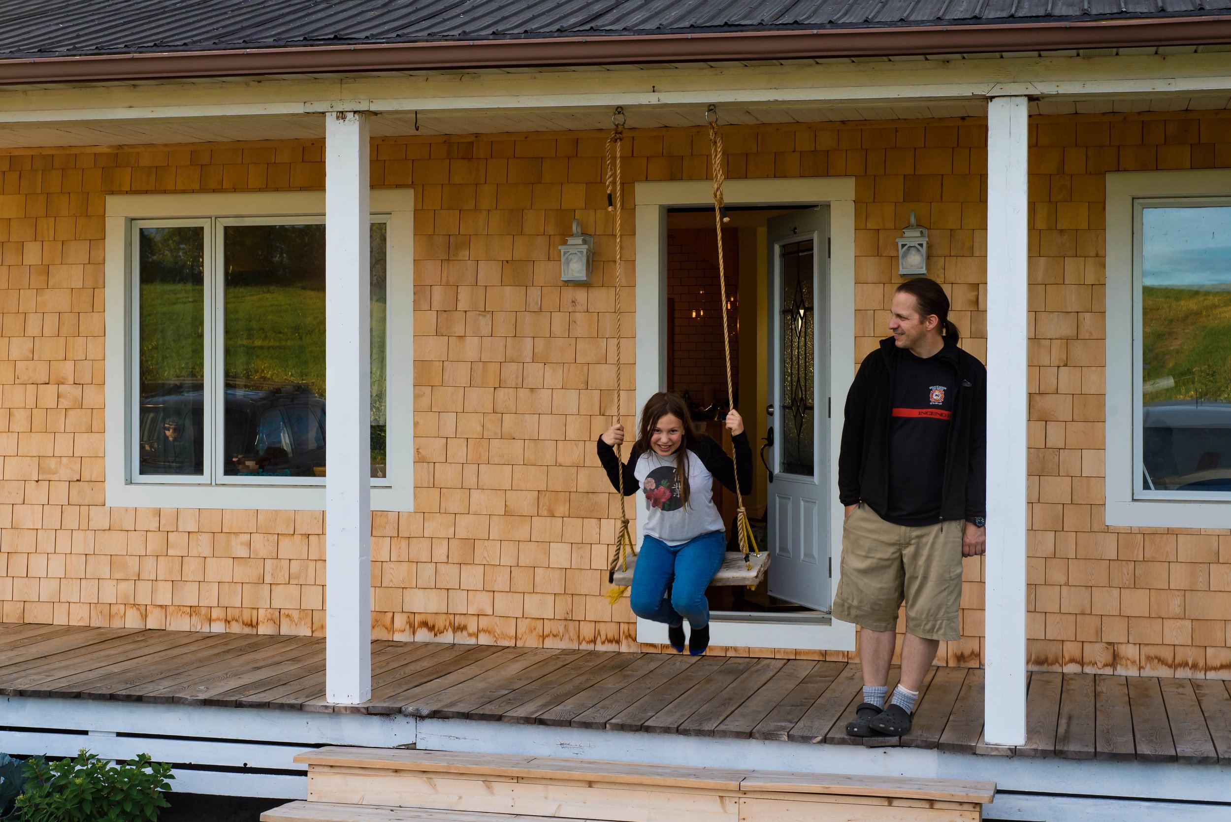 A girl sitting on a porch swing with her father standing next to her in front of a country house with wooden siding.