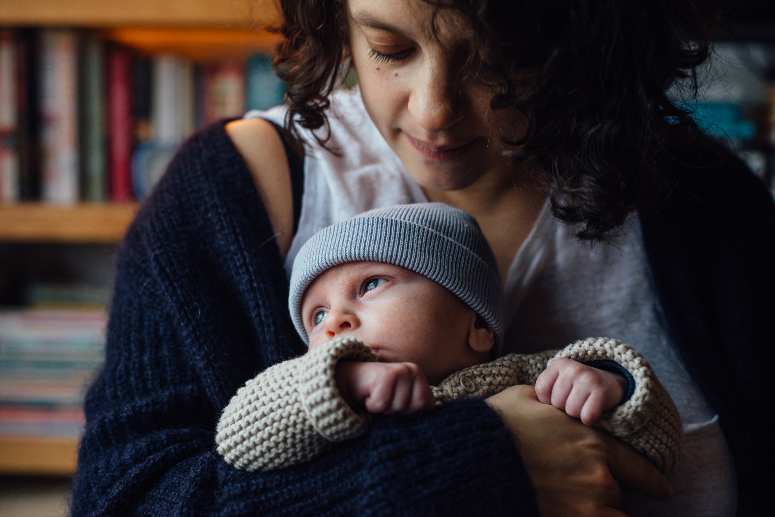 A woman with curly hair and closed eyes looking down at a baby in her arms, who is wearing a gray beanie and a cream-colored knit sweater, inside a home with a bookshelf in the background.