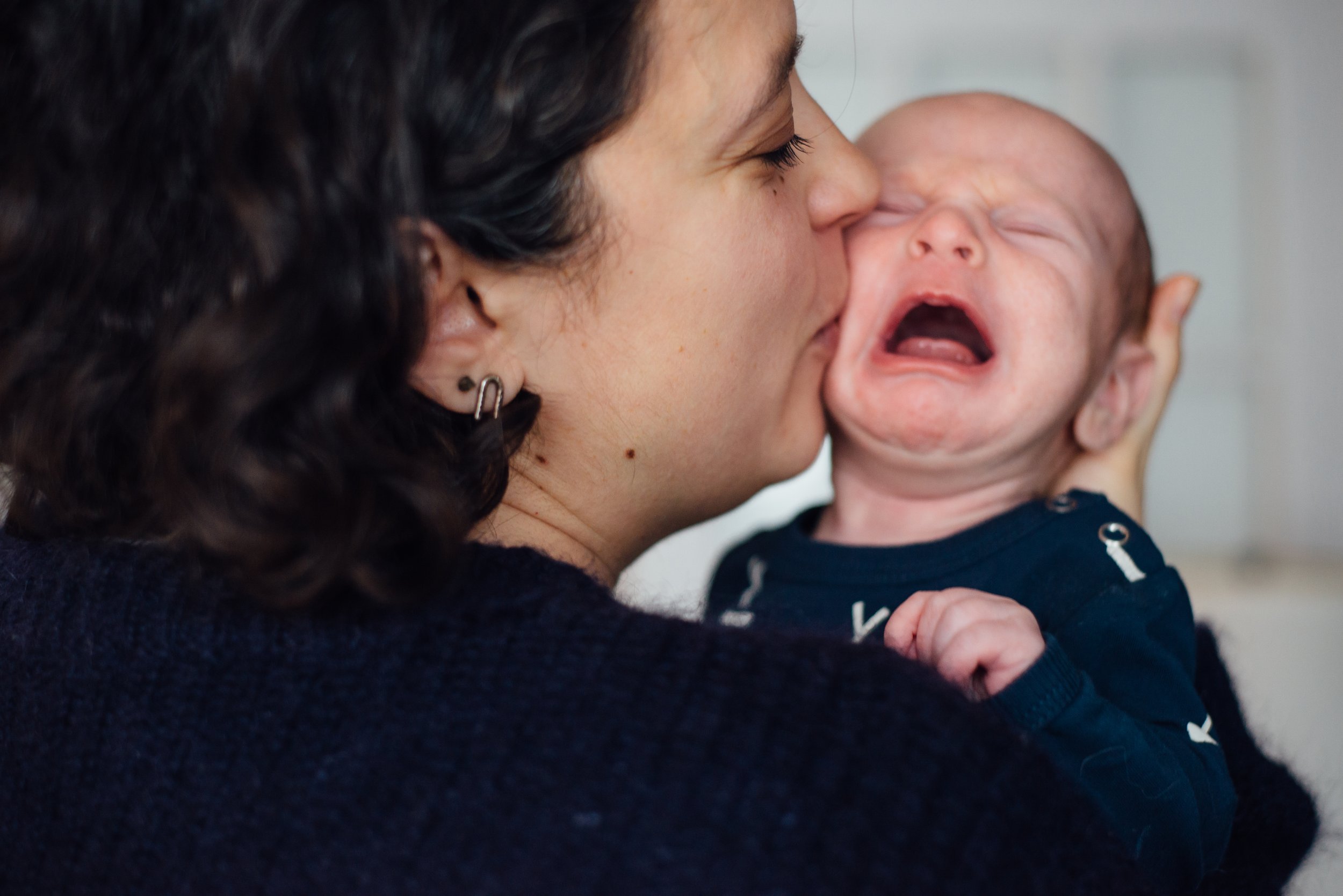 A woman with dark curly hair and earrings holding a crying baby close to her face.