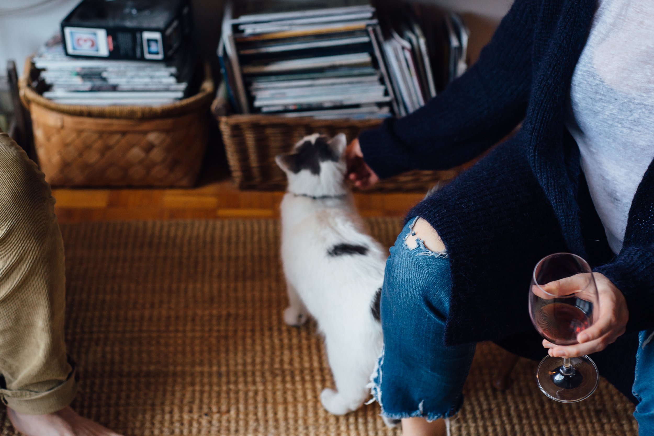 A person holding a glass of red wine, sitting on a chair or sofa, with a black and white cat sitting on the floor nearby. The scene is indoors, with woven baskets and shelves filled with vinyl records in the background.