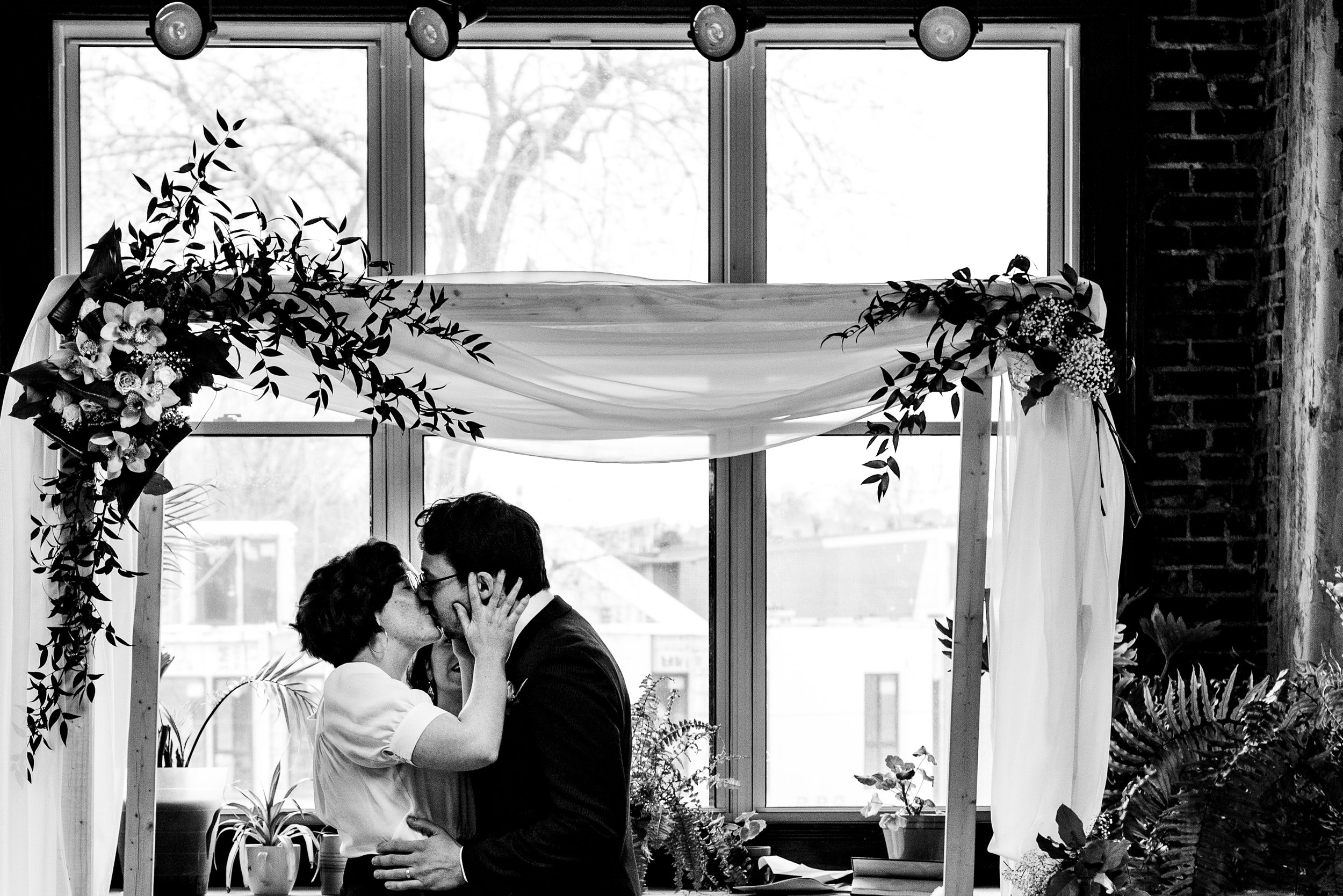 Black and white portrait of Bride and groom exchanging first kiss in front of wedding altar.