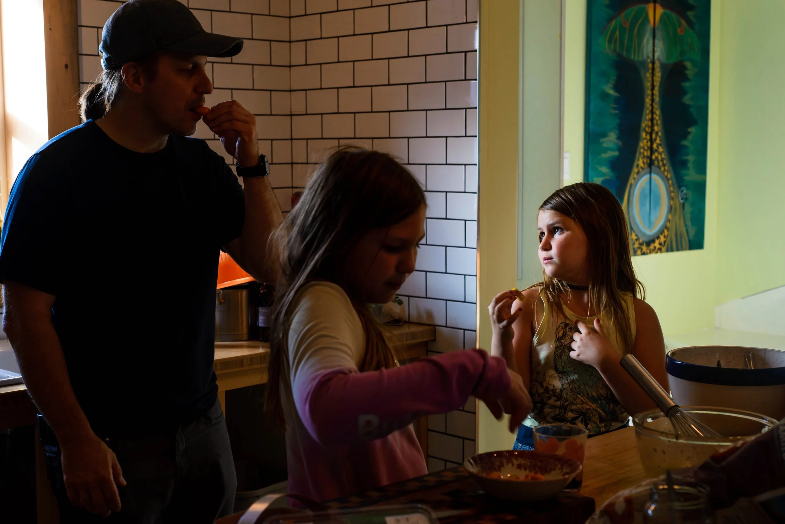A man wearing a cap and Black T-shirt watching two young girls baking in a kitchen. One girl is stirring a mixture in a bowl, while the other girl is looking at her and eating a carrot.