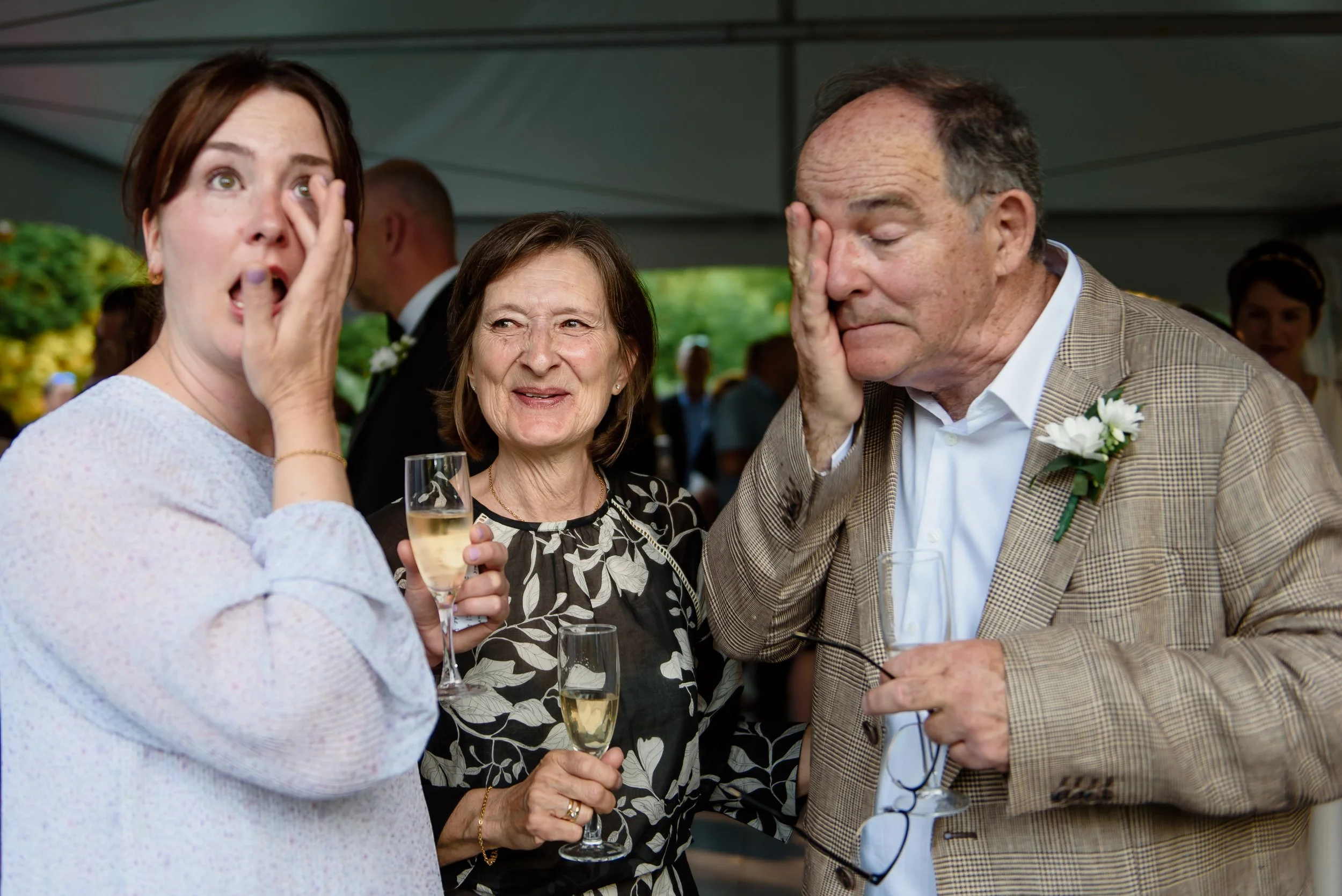 Three people stand together at an event, two holding champagne glasses. One woman and one man wipe their eyes, while another woman smiles in the centre