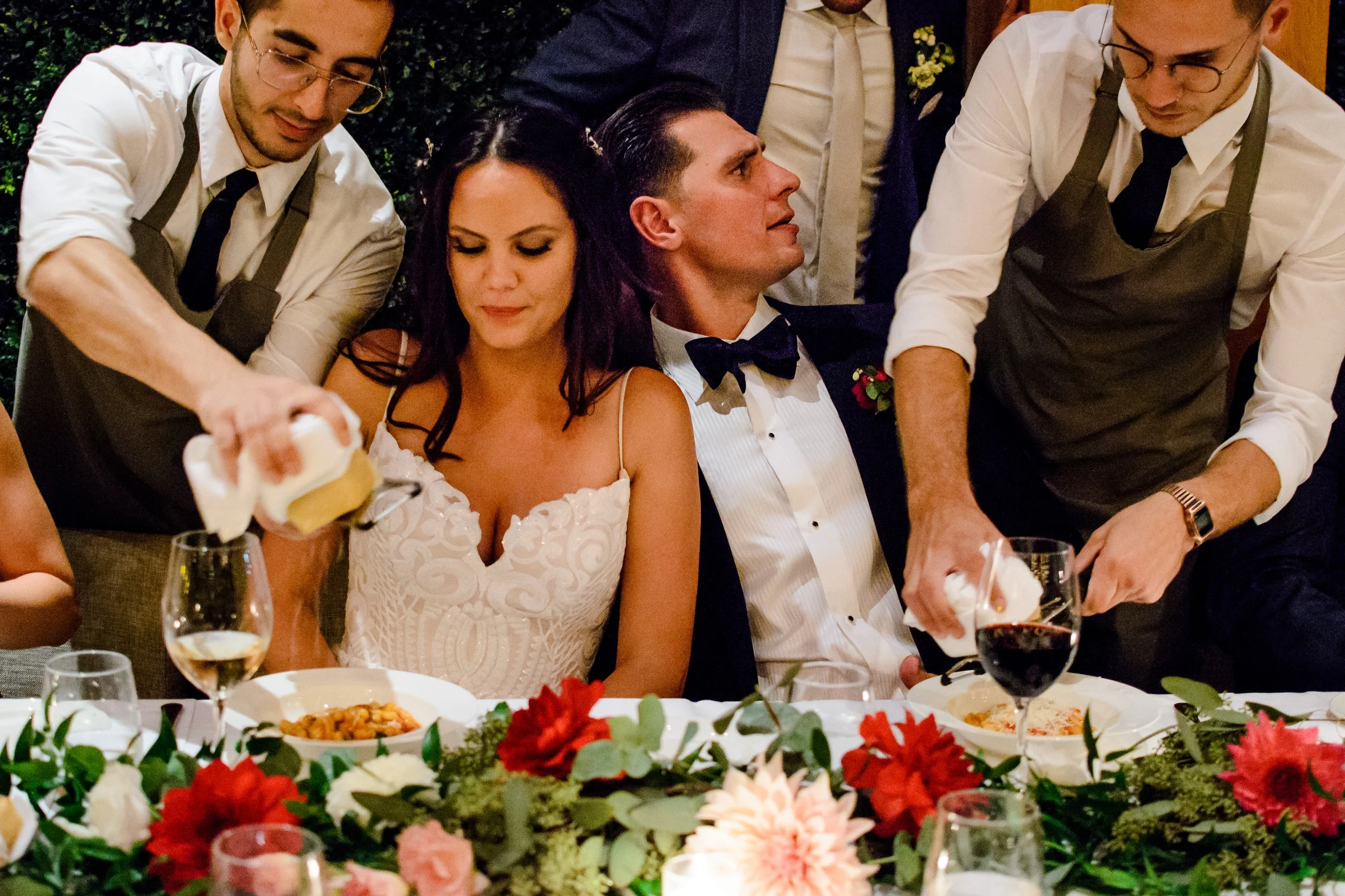 A group of people sitting at a table during a wedding reception. The bride and groom are in the center, with the bride wearing a white dress and the groom in a tuxedo with a bow tie. They are surrounded by waitstaff serving drinks. The table is decorated with a colorful floral centerpiece and food dishes.