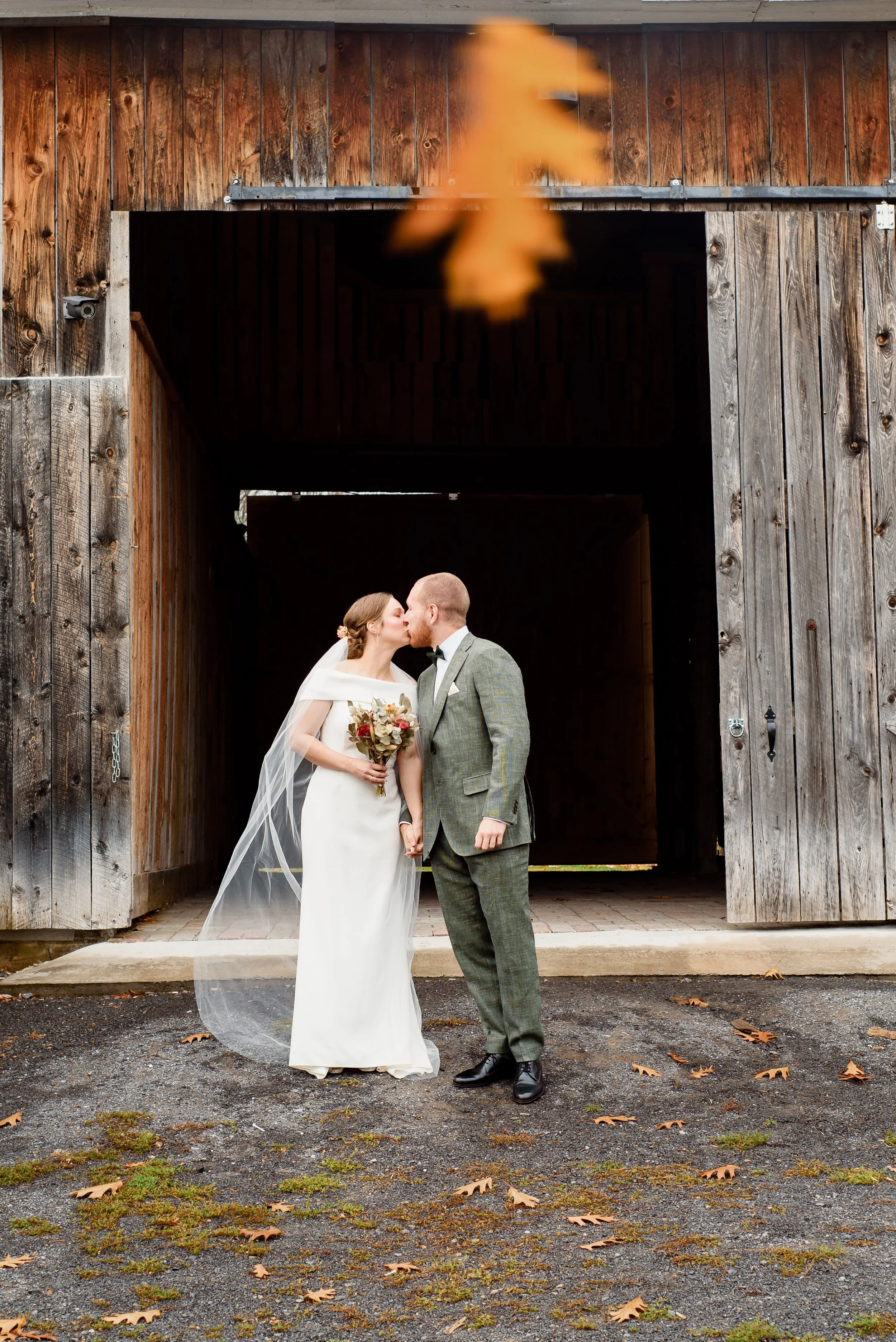 A bride and groom kissing in front of a rustic wooden barn during their Lanaudière wedding, with fallen leaves on the ground.