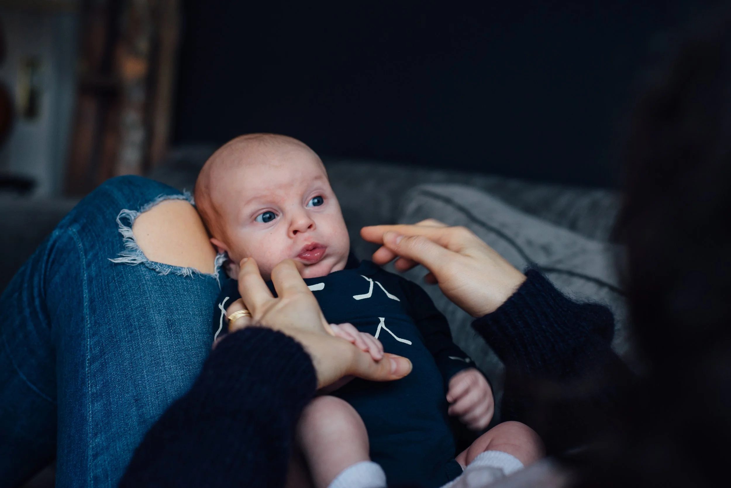 A baby with light skin and blue eyes lying on an adult's lap, looking up and touching their face, while the adult gently holds the baby's chin. The scene appears to be indoors.