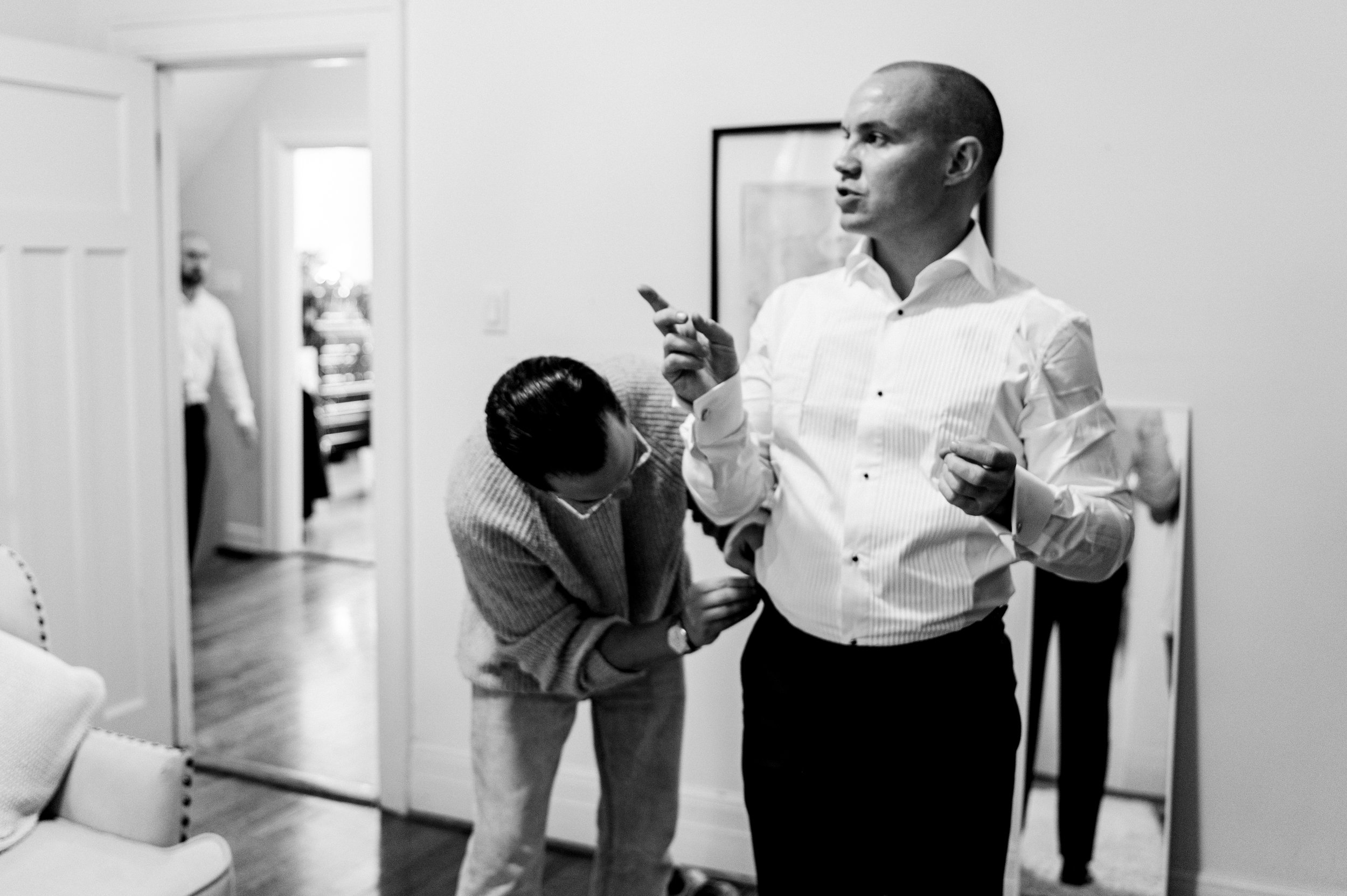 Groom stands and points as he talks  to someone off camera while groomsman works on his suspenders