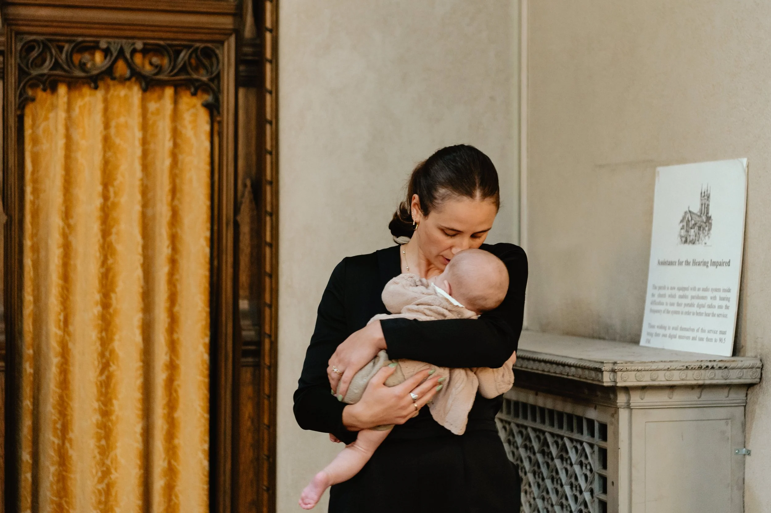 Wedding guest rocks baby in front of Montreal church confessional with yellow drapes