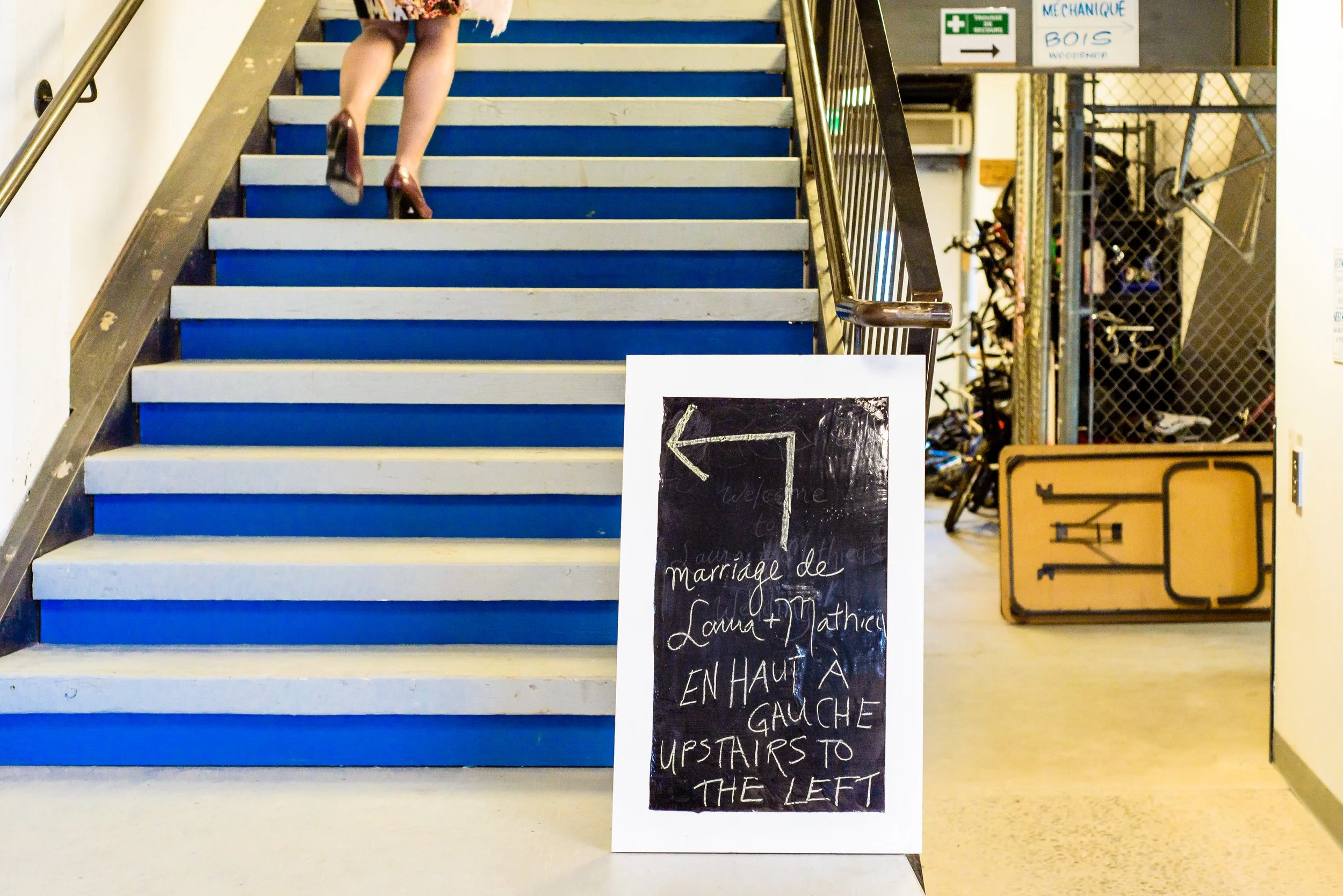 a sign announcing the wedding of Laura and Matthieu up and to the left sits in front of white and vivid blue stairs. On the top left of the frame we see the legs of a person running up the stairs.