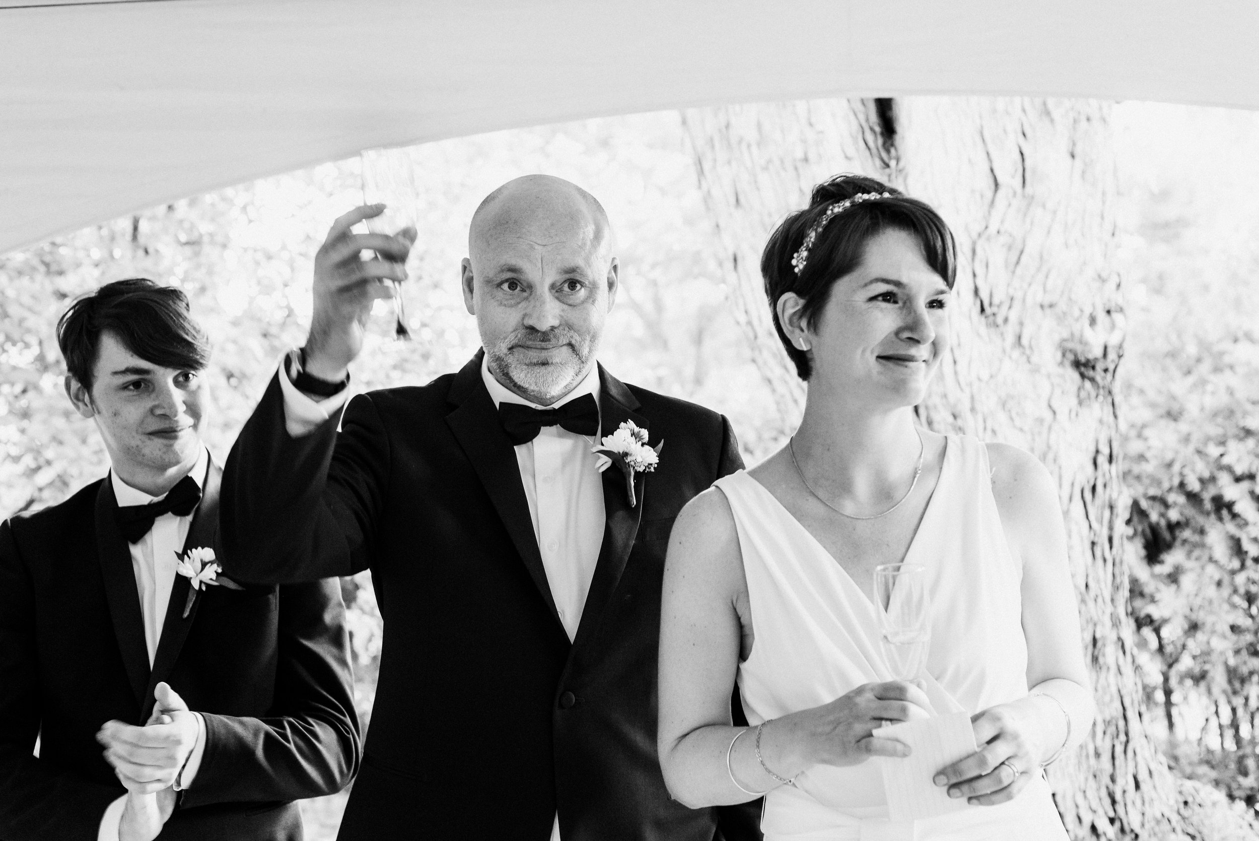Three people stand together at wedding, two men wearing tuxedos and a woman wearing a white bridal pantsuit. The groom raises his glass, the bride hold a piece of paper and a young man applauds.