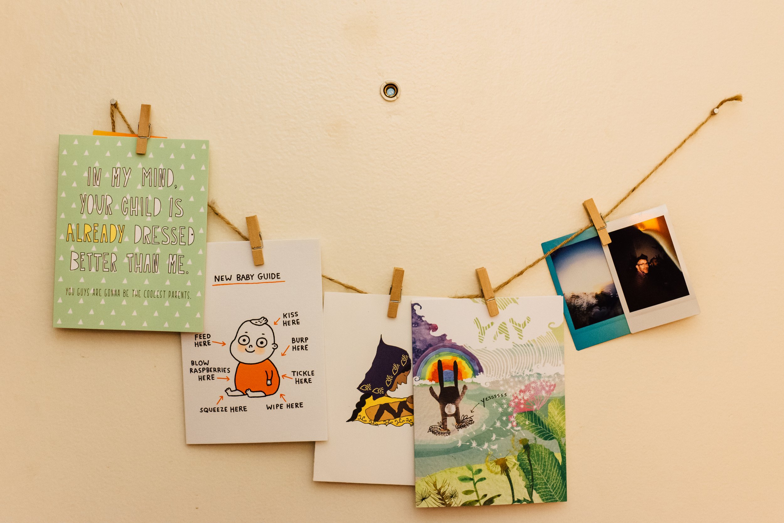 A collection of baby congratulations cards and photographs hanging on a string with small clothespins, displayed on a door. Above the string we see the door's peephole.