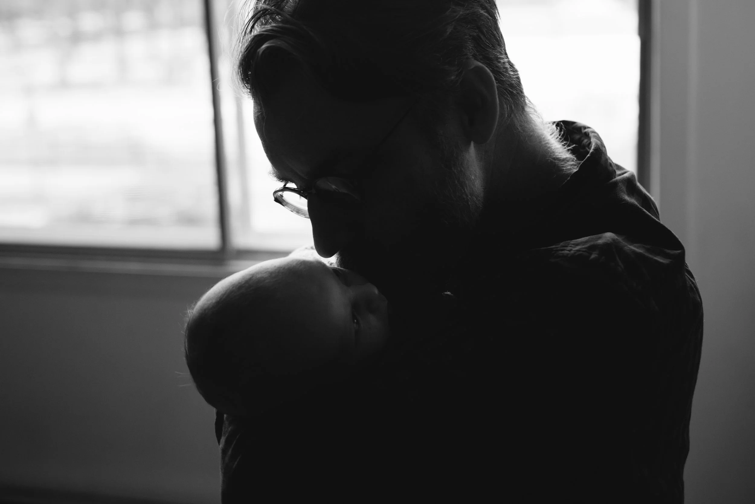 A black and white photo of a person with glasses and a beard, bending over a newborn baby's head. The person appears to be comforting or caring for the child near a window with bright light coming through.