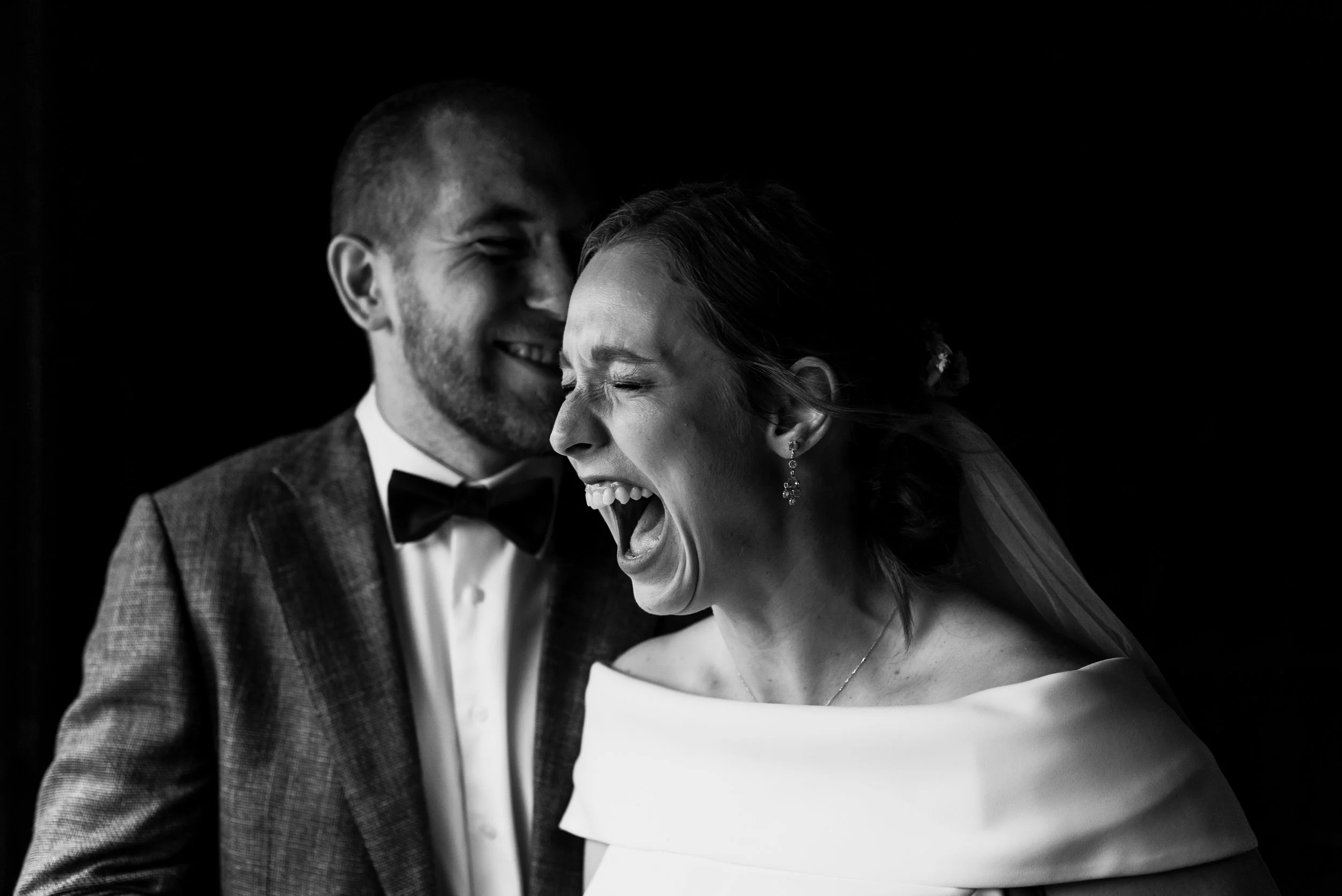 Black and white photo of a groom and bride laughing and sharing a moment of joy at their wedding, with the groom in a tuxedo and the bride in a wedding dress.