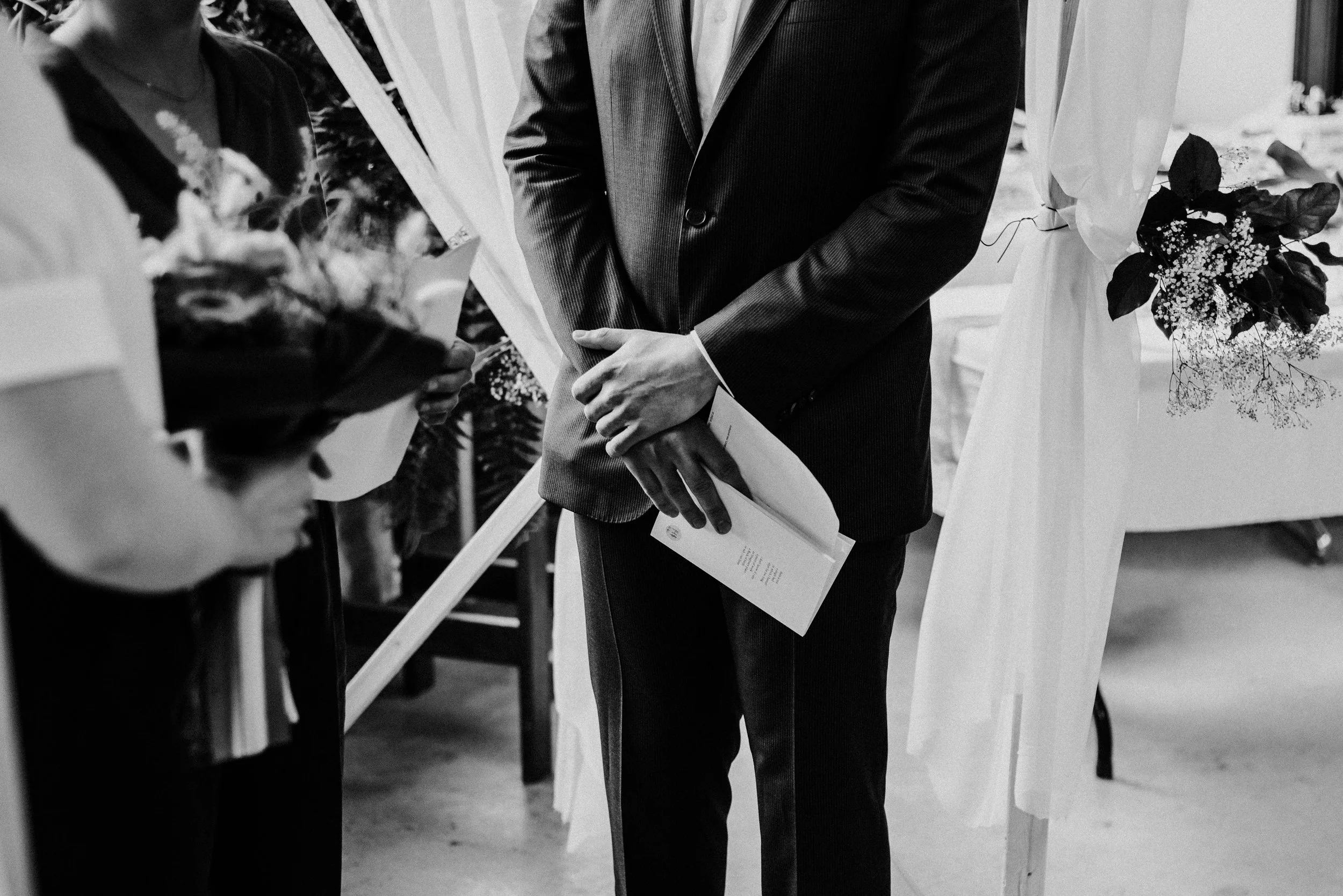 Black and white close-up of a bride and groom at the altar, focusing on the groom's hands holding written vows.