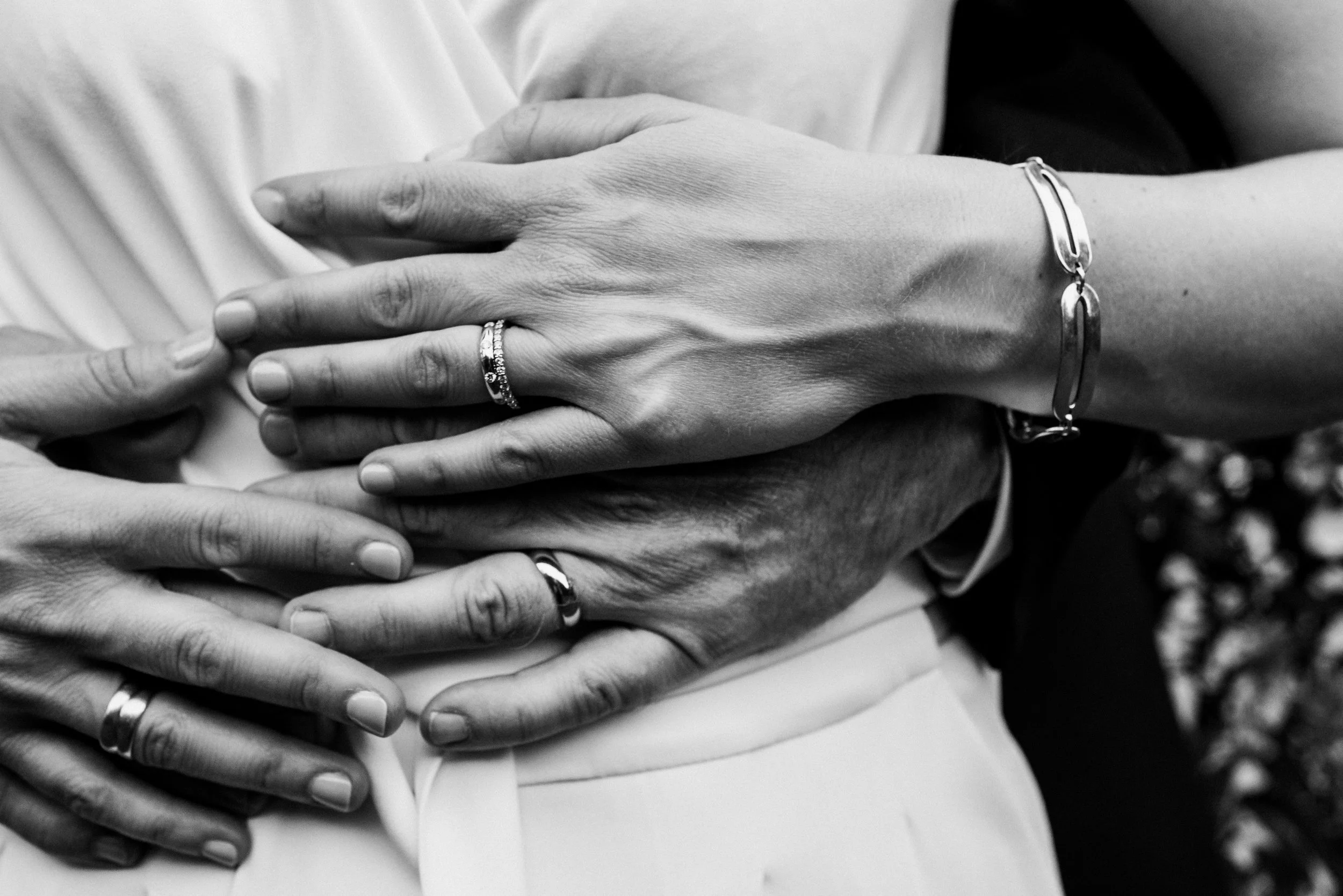 Close-up of two people embracing, each wearing wedding rings, with their hands overlapping against a blurred background.