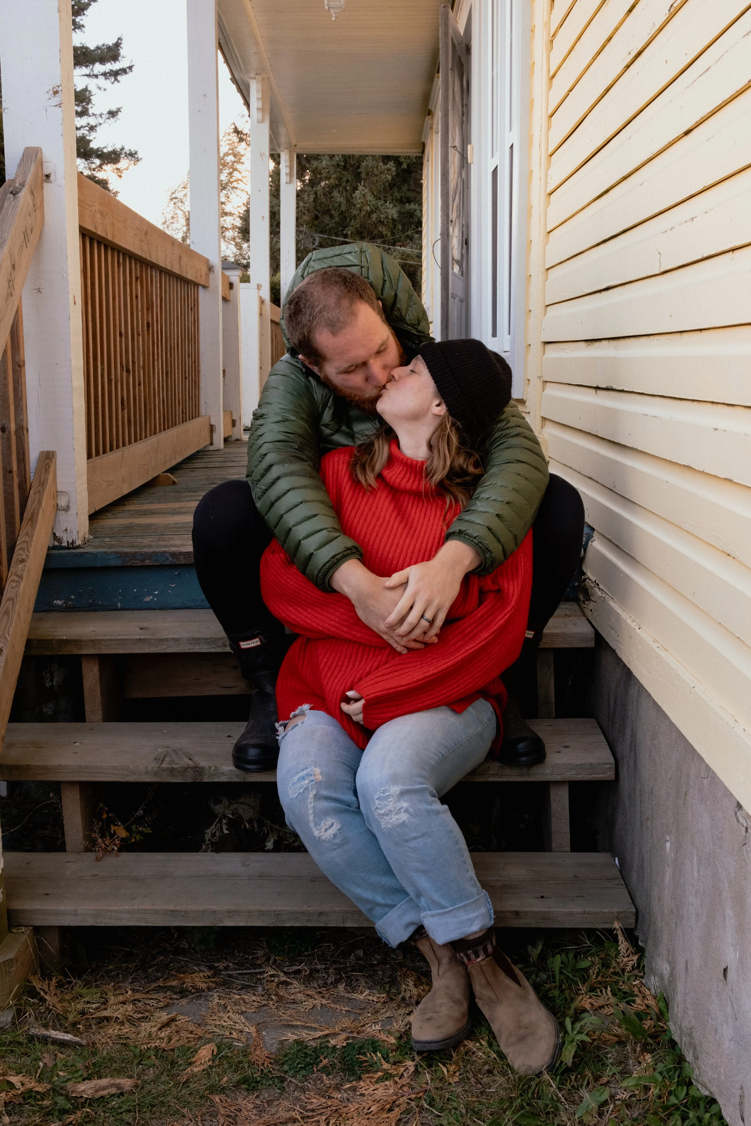 A man and woman share a kiss on the porch steps of a yellow house, with the woman sitting on a step and the man leaning over her. The woman wears a black knit hat and red sweater, the man wears a green jacket and has a backpack. They are outdoors, wi