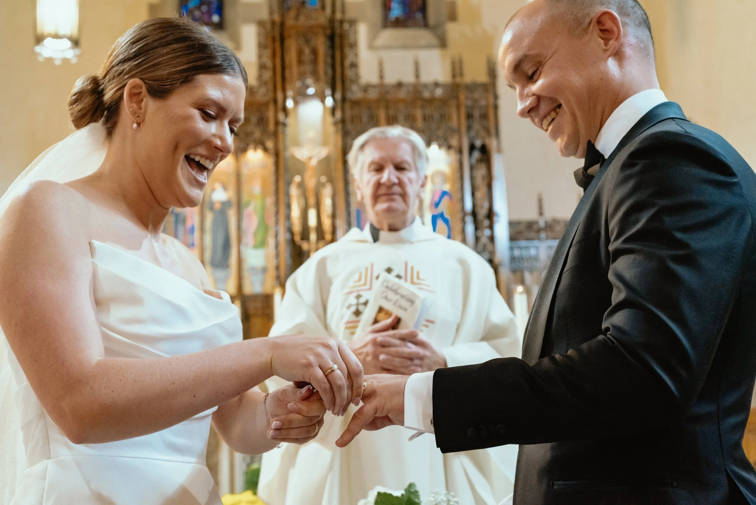 Groom and Bride smile widely as bride places wedding band onto groom's finger and Montreal pastor watches