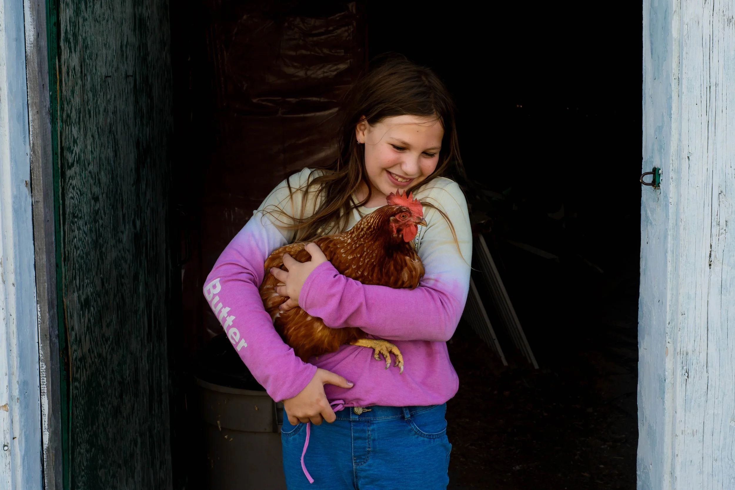 A young girl with long brown hair, wearing a pink long-sleeve shirt, holding a brown hen, standing in a doorway with weathered blue and white wooden walls.