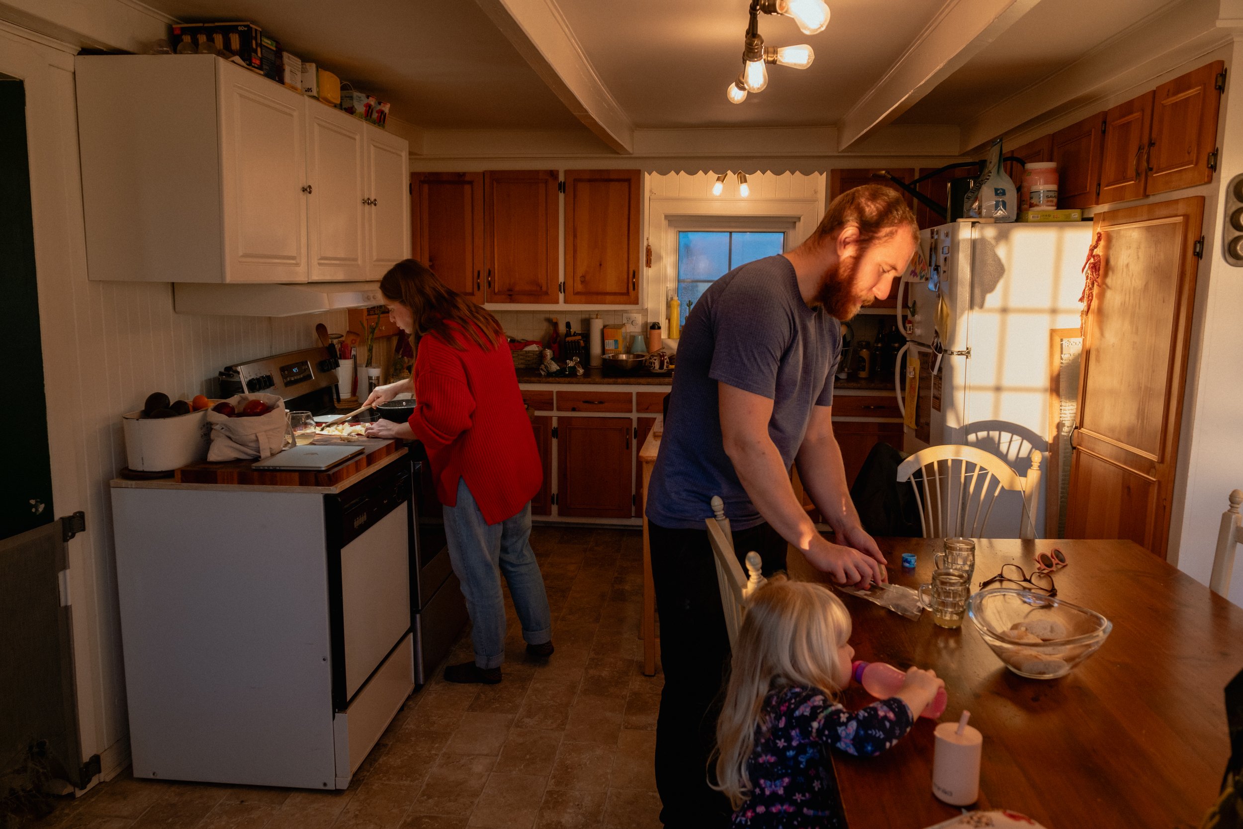 A family in a kitchen preparing a meal together. There is a man and a woman working at the table and counter, and a young child seated at the table. The kitchen has wooden cabinets and a window with warm evening sunlight streaming in.
