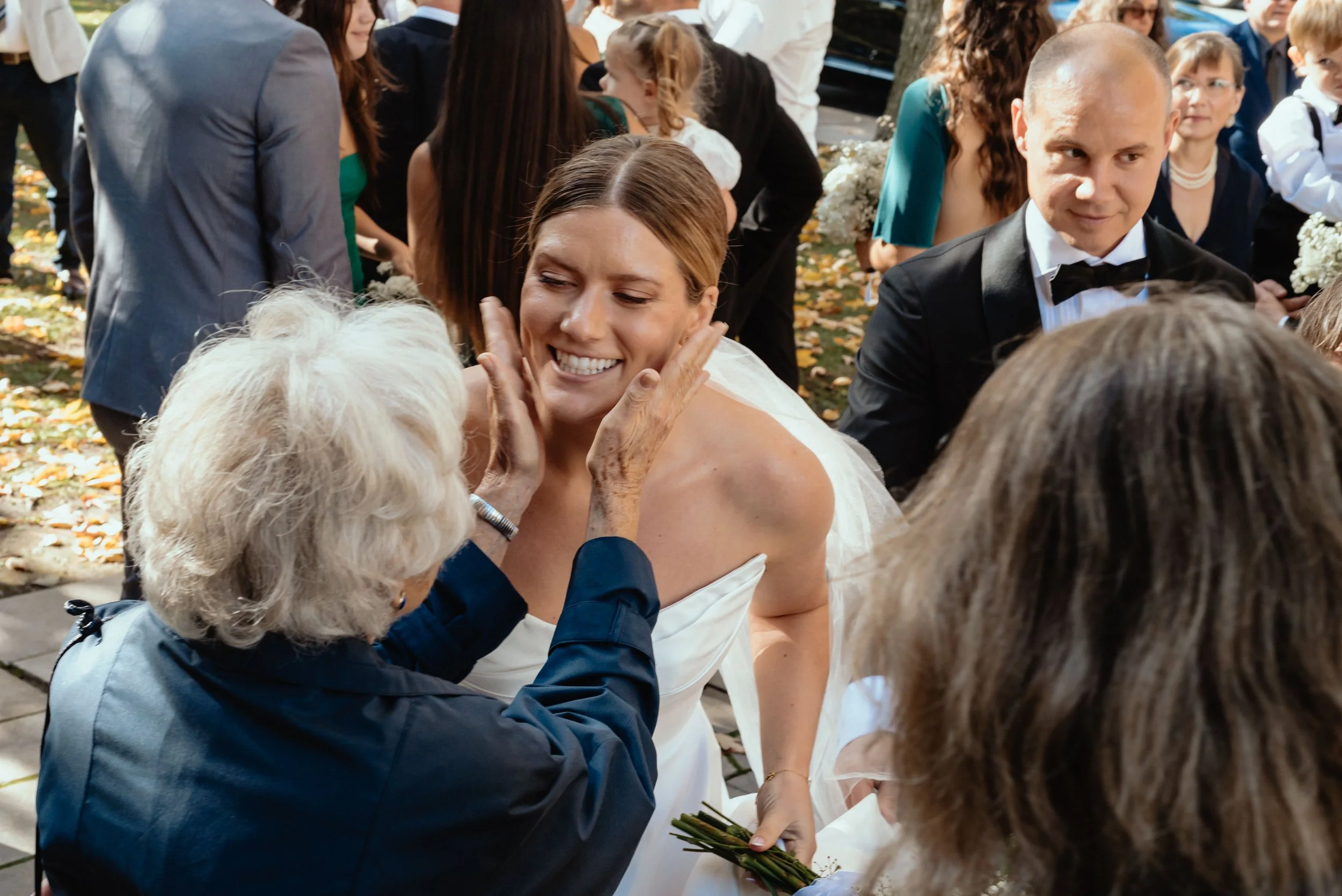 Montreal bride and groom surrounded by wedding guests as grandmother joyfully clasps the bride's cheeks in congratulations.
