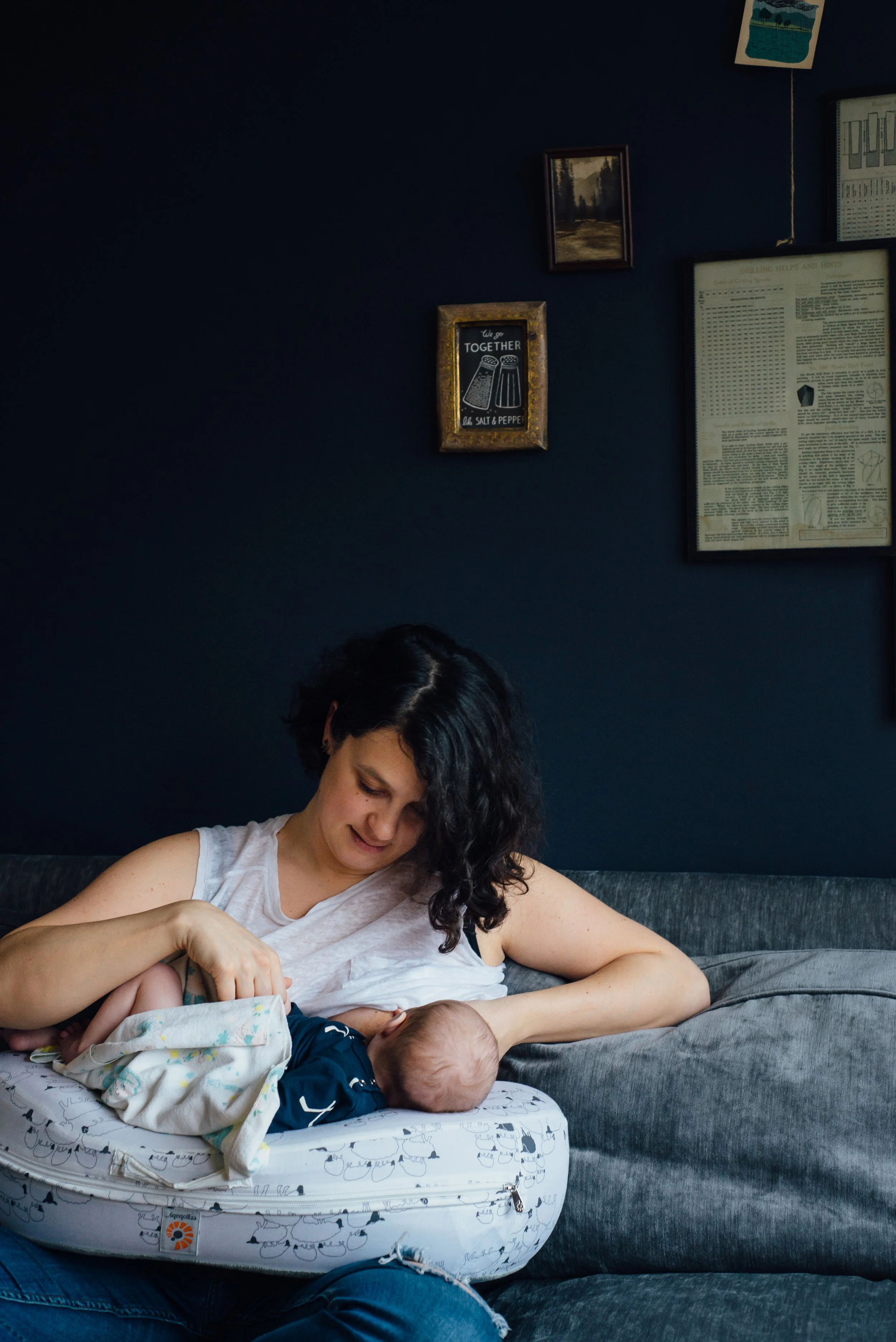 A woman with curly dark hair, wearing a sleeveless white shirt, sitting on a gray couch, tending to a baby lying on a white baby mattress with a pattern of animals and trees in a room with dark blue walls, decorated with various framed pictures and p